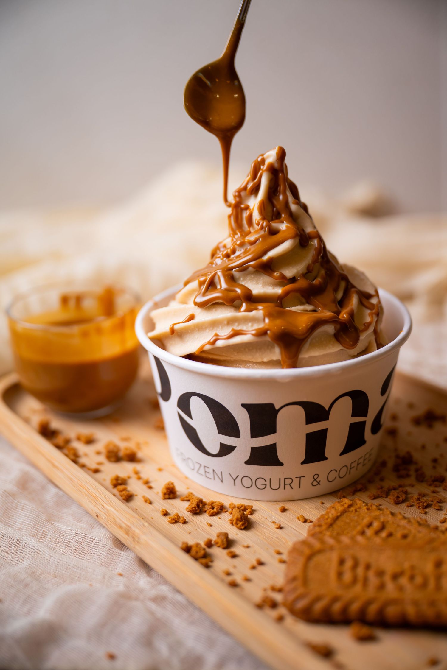 Caramel sauce drizzled over swirled frozen yogurt in a Poma cup, served on a wooden tray with a Biscoff cookie.
