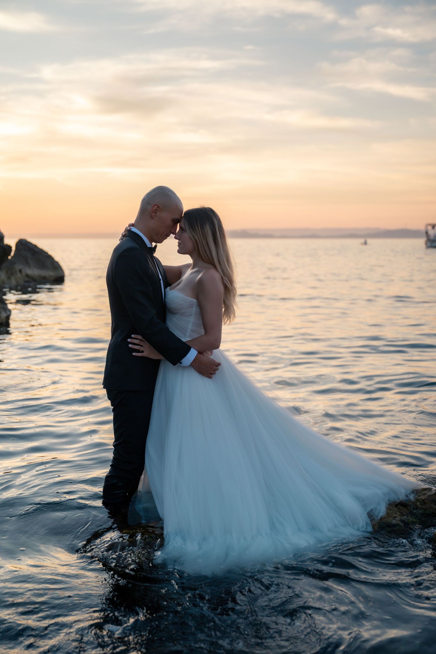 Newlyweds embrace forehead-to-forehead standing in shallow ocean water at sunset.