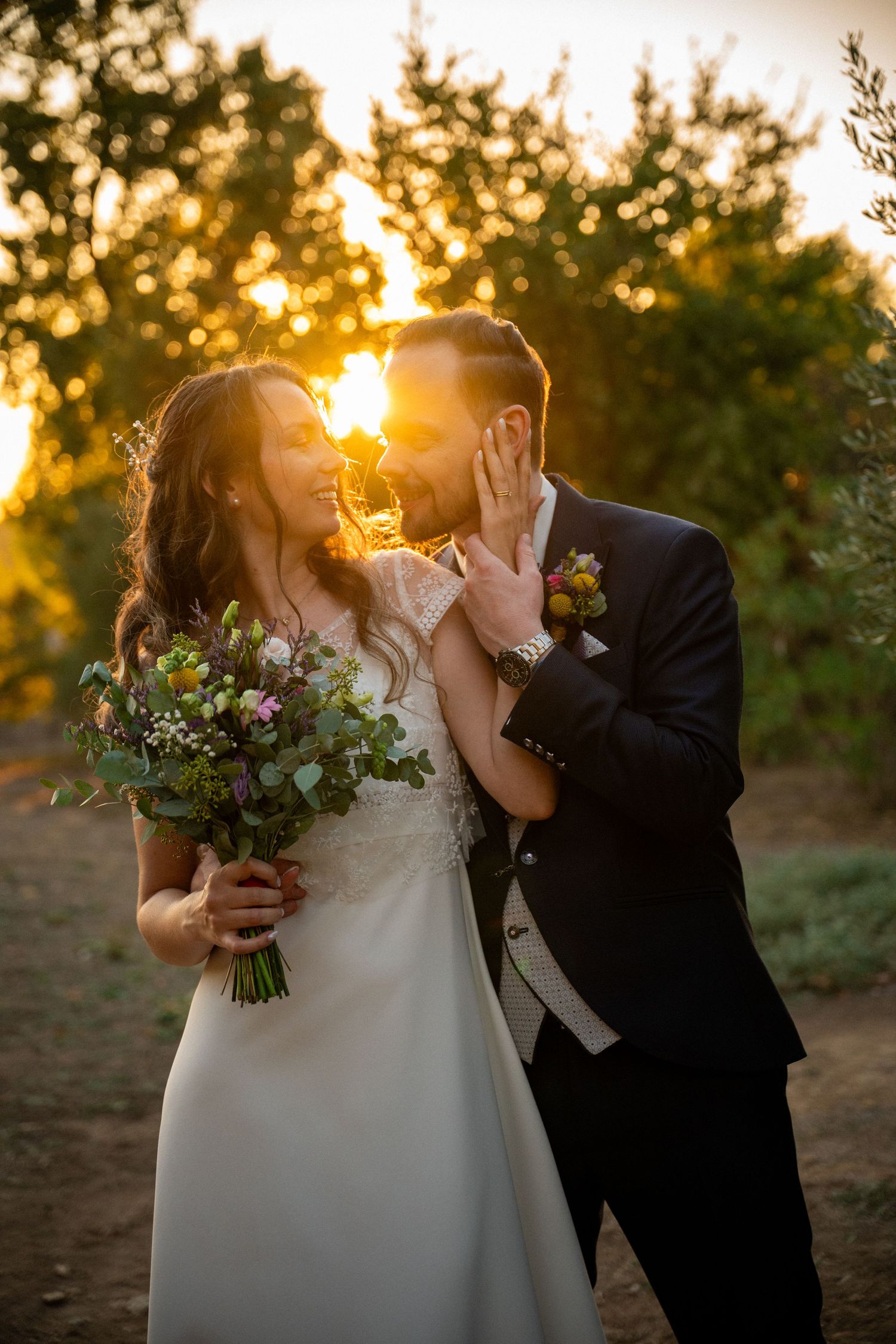 Bride and groom kissing at sunset in an olive grove, bride holding wildflower bouquet.