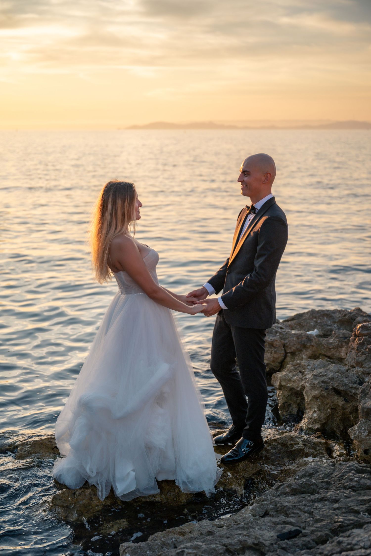 Bride and groom hold hands on rocky shore at sunset, ocean and mountains in background.