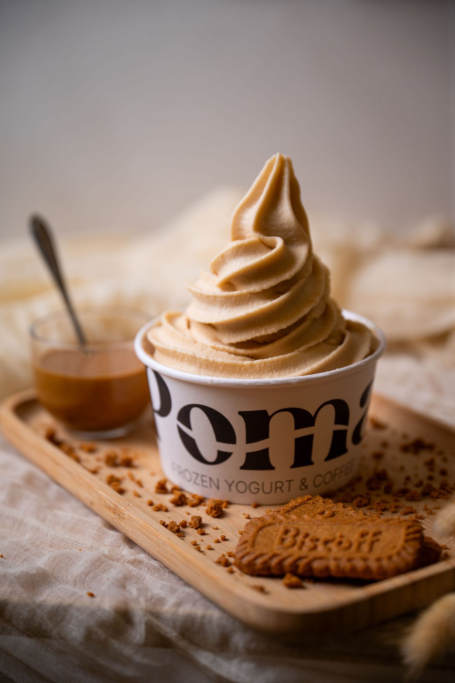 Swirled caramel soft-serve frozen yogurt in a paper cup on a wooden tray with a Biscoff cookie and caramel sauce.