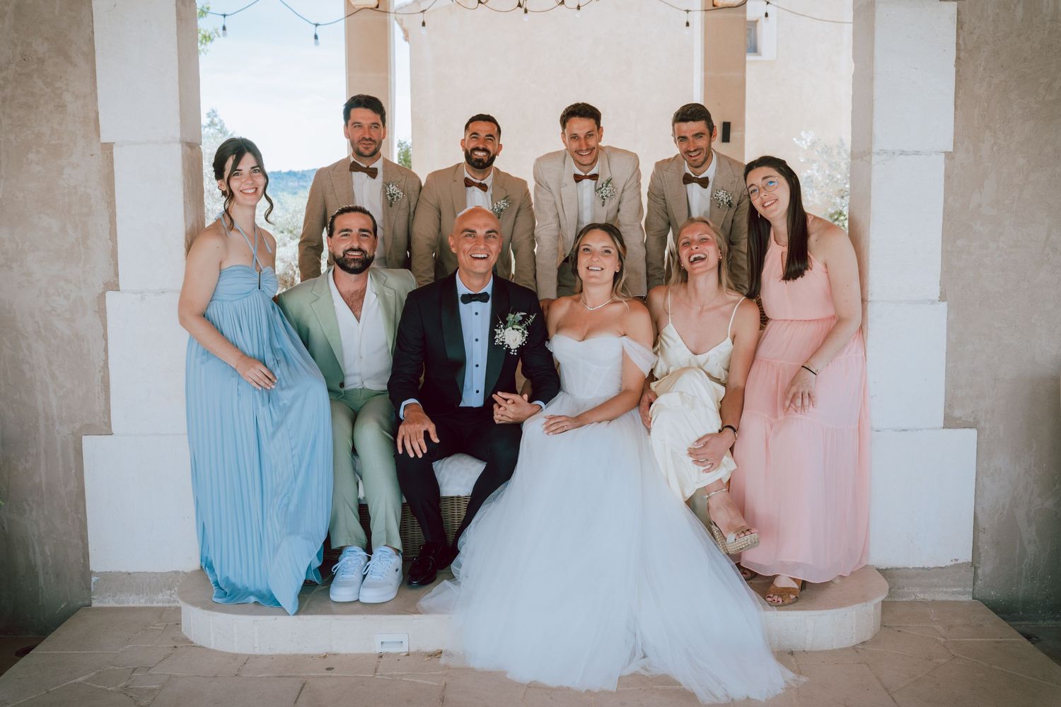 Wedding party group photo with bride, groom, bridesmaids and groomsmen posing together at a sunny venue.