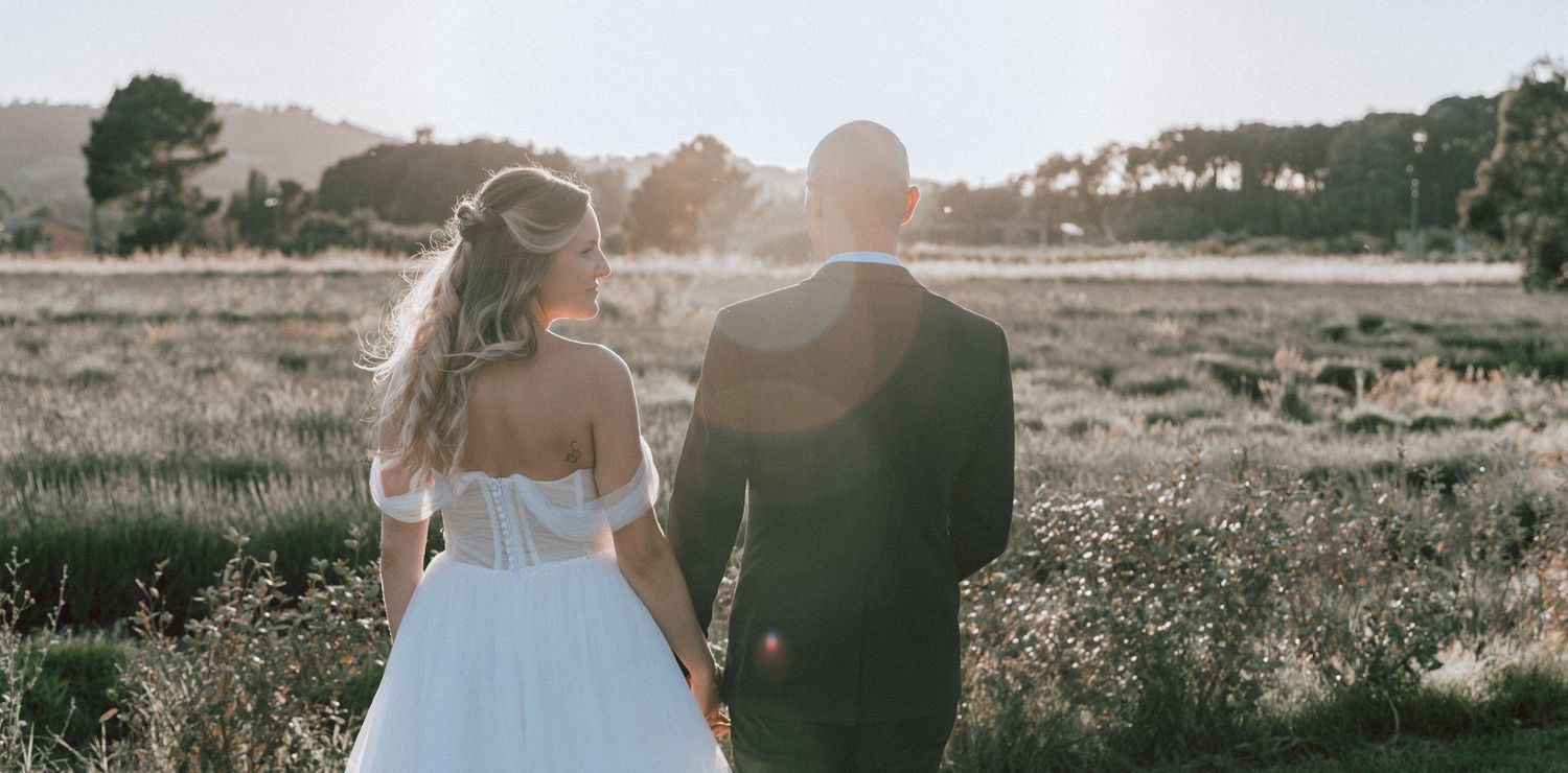 Bride and groom hold hands walking through a golden sunlit meadow during their wedding day.