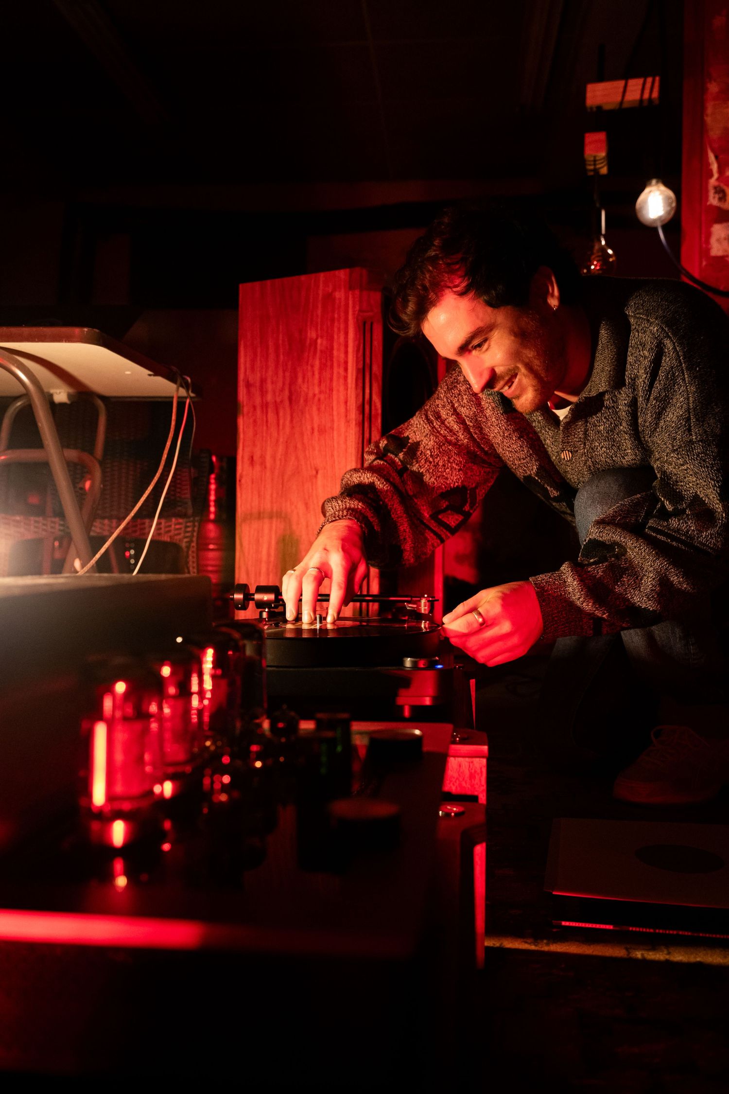Man smiling while operating a DJ turntable setup under warm red ambient lighting in a dimly lit room.