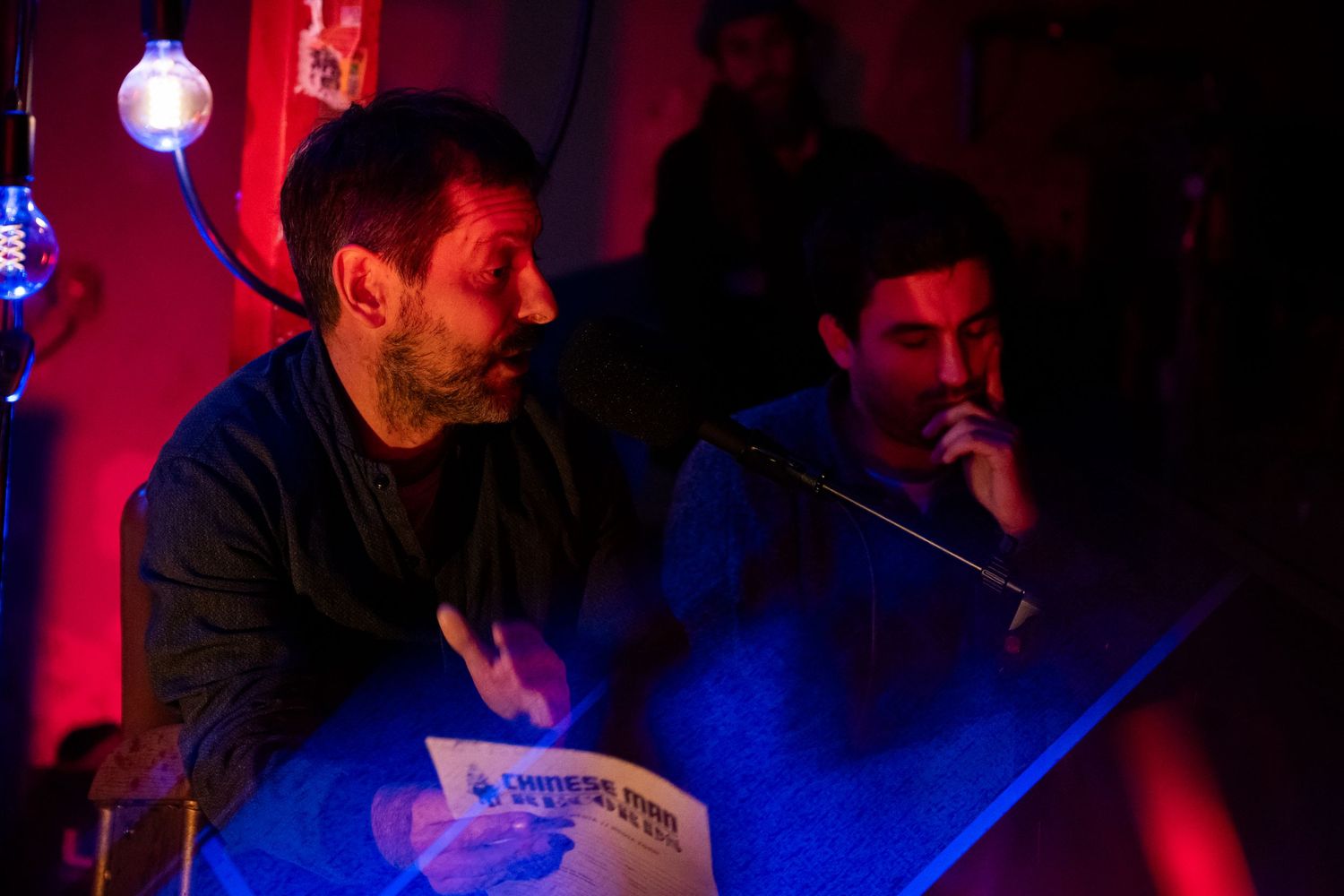 Two men sit at a table in a dimly lit bar with red and blue lighting, one holding papers.