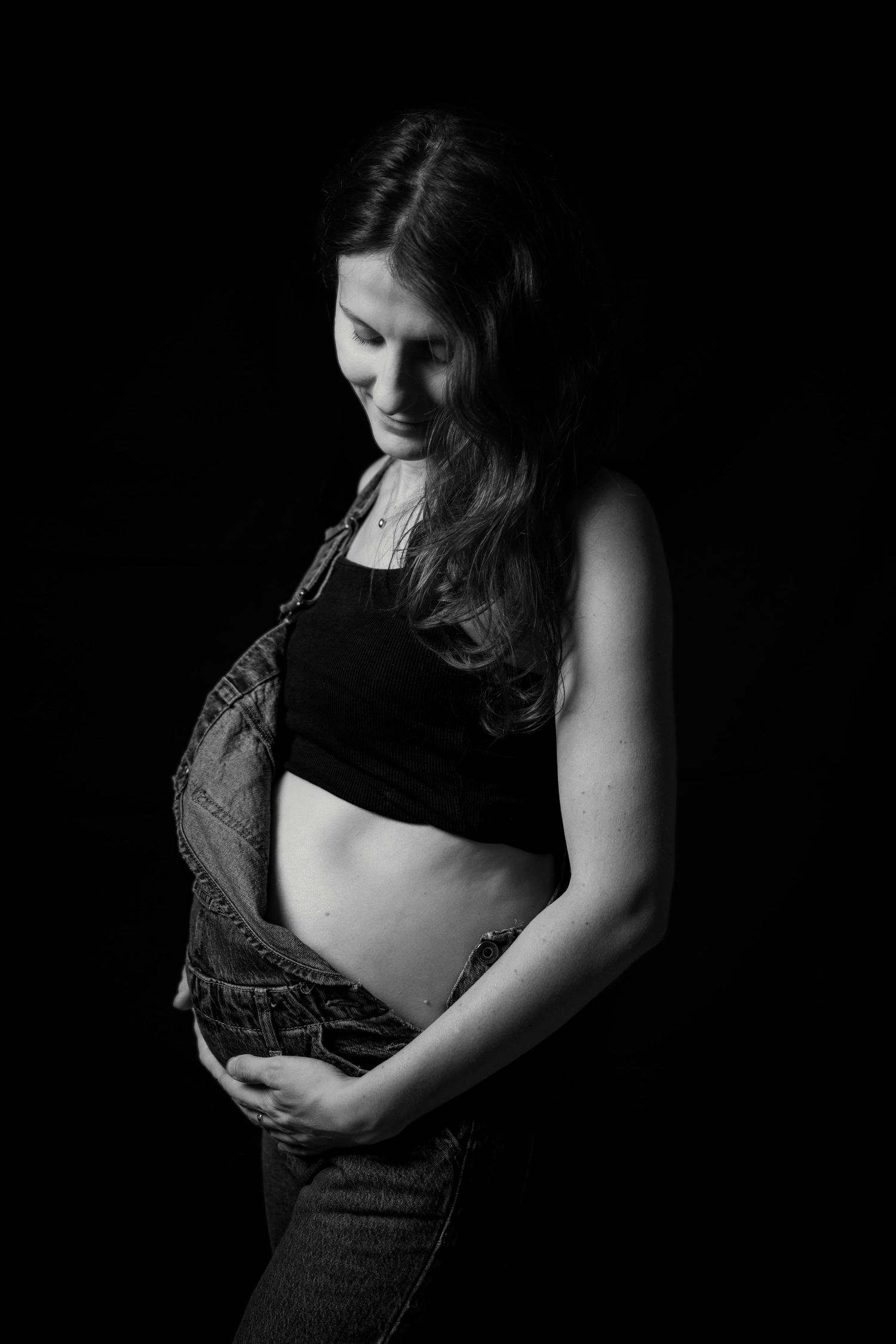 Pregnant woman in denim overalls and black top gently holding baby bump in black and white studio portrait.