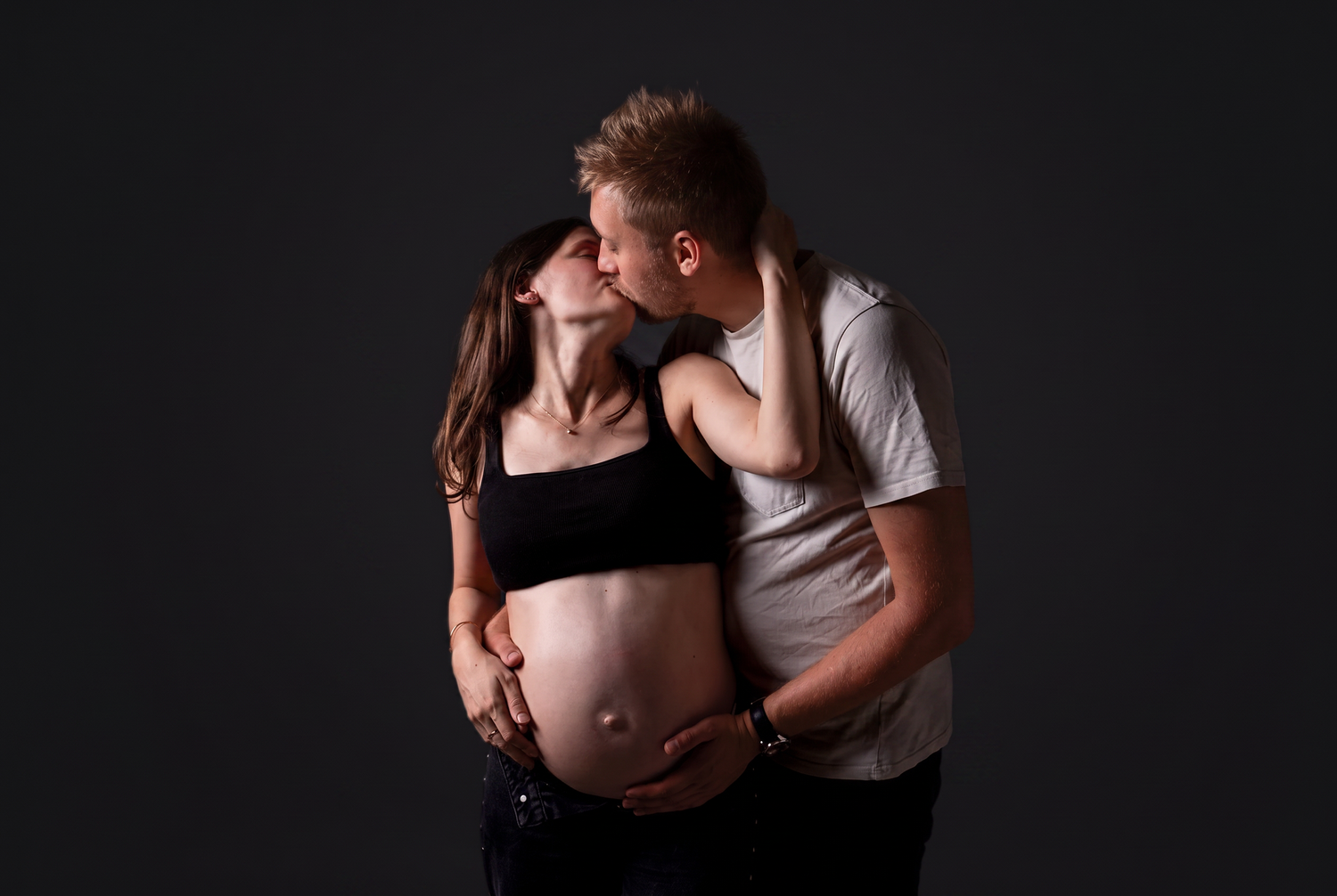 Expecting couple kissing in dark studio, man embracing pregnant woman in black crop top and jeans from behind.
