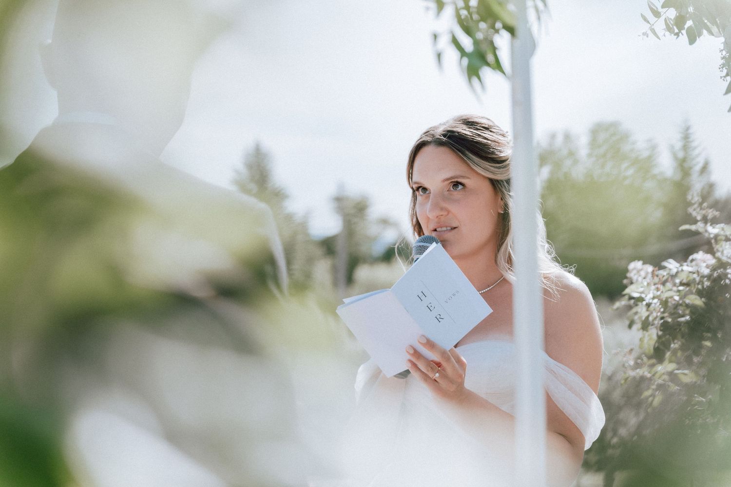 Bride in white gown reads vows from a card during an outdoor wedding ceremony.