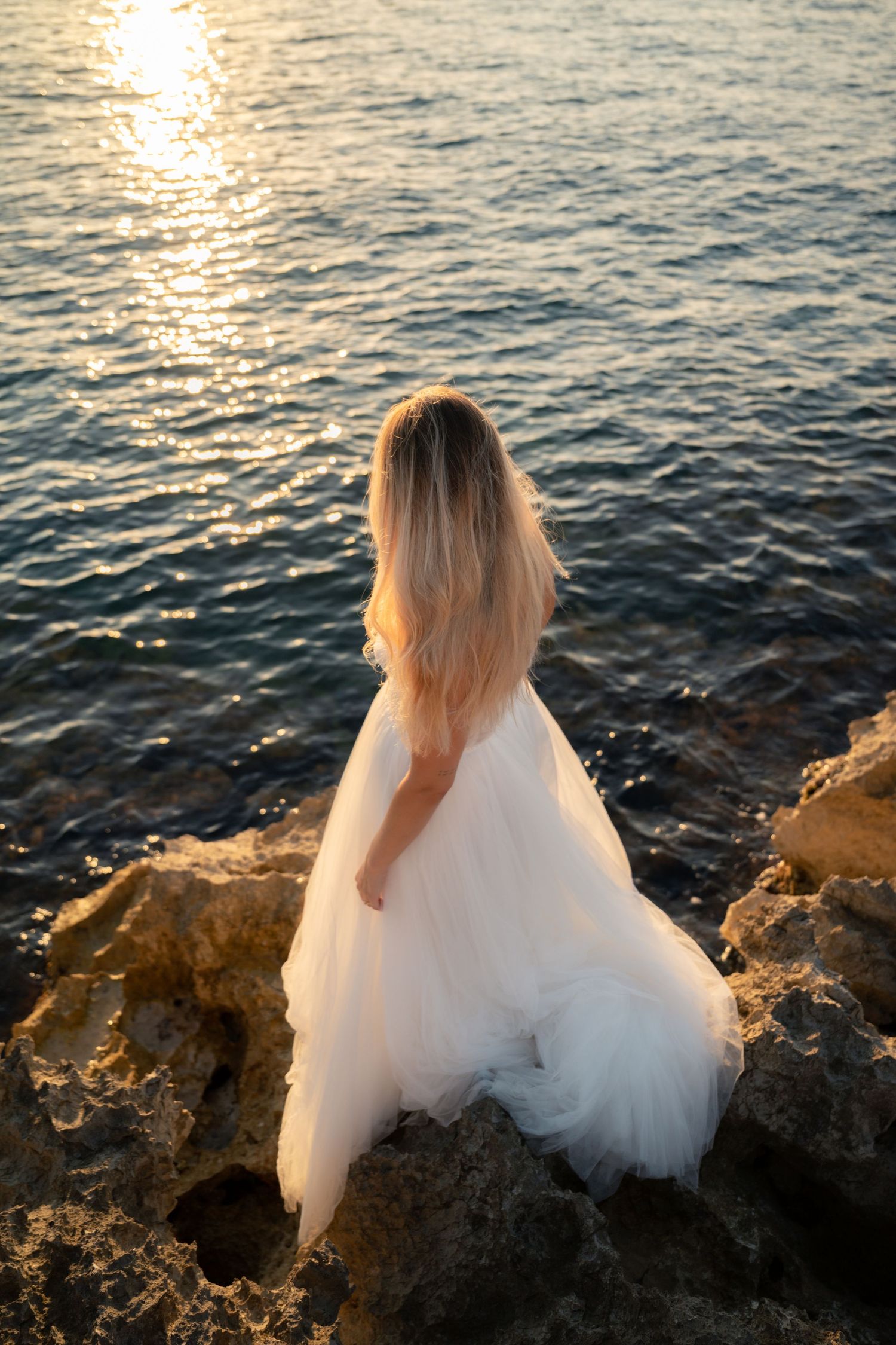Bride in white tulle gown sitting on rocky shore gazing at golden sunset reflecting on calm ocean waters.