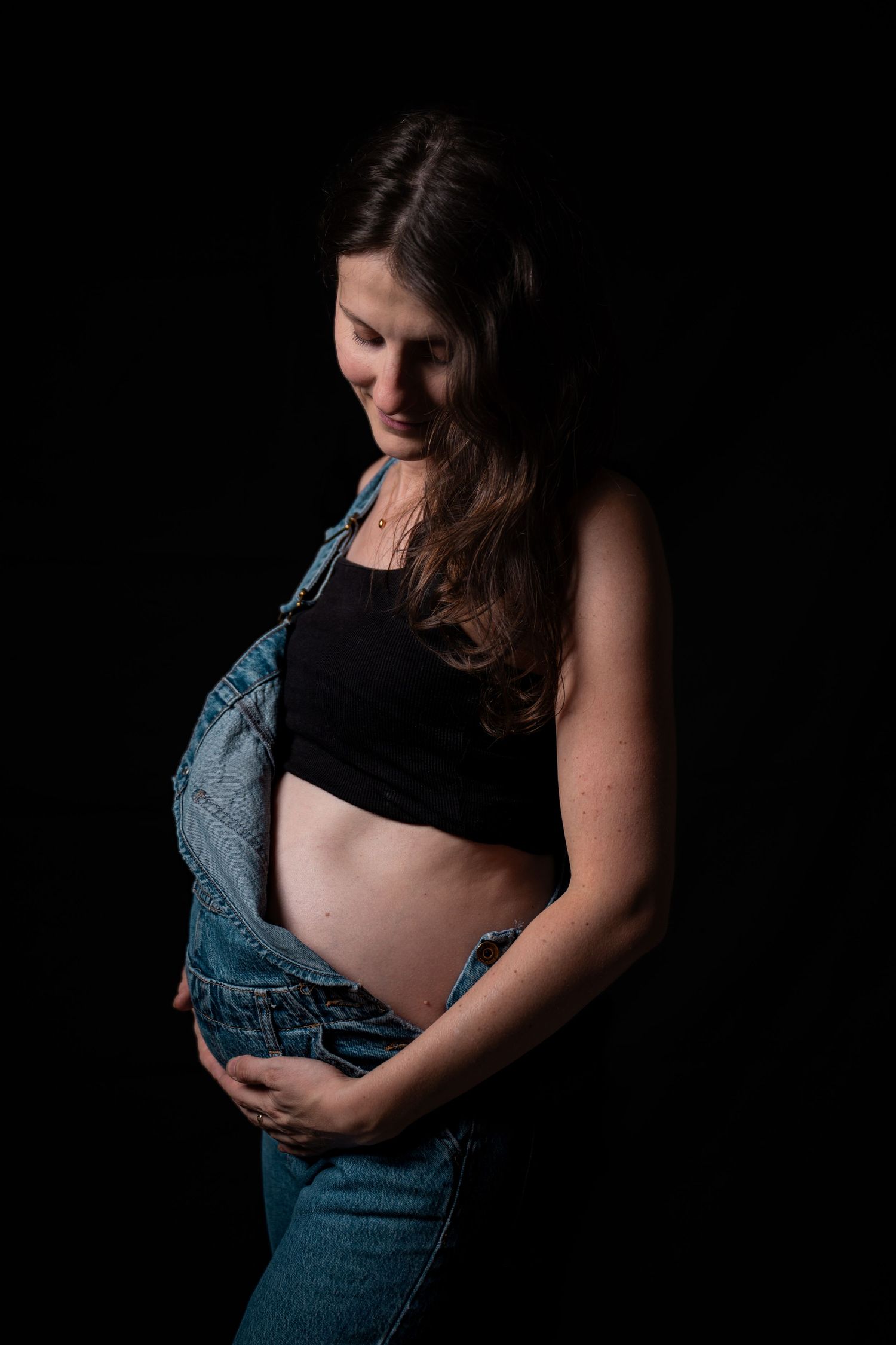 Pregnant woman in denim overalls and black crop top cradling her baby bump against a dark background.