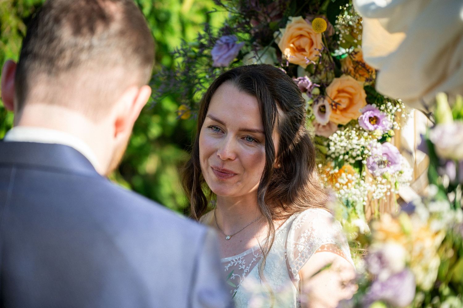 Bride gazing emotionally at groom during outdoor wedding ceremony with colorful floral arch backdrop.