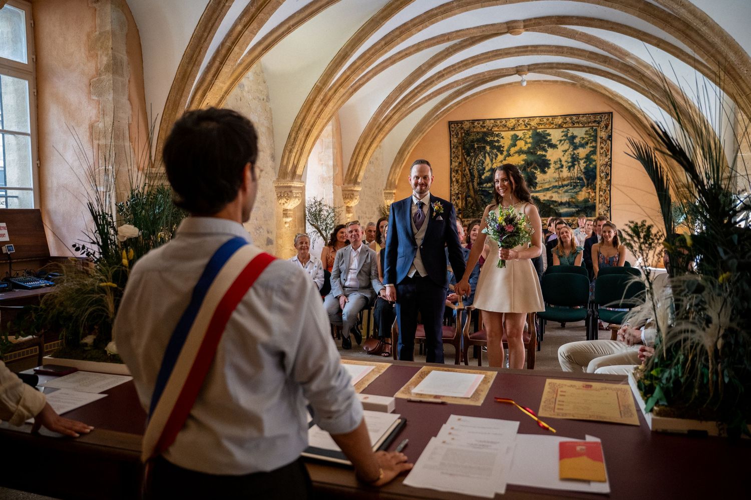 Couple walks down aisle in elegant vaulted stone hall during French civil wedding ceremony with guests seated behind.