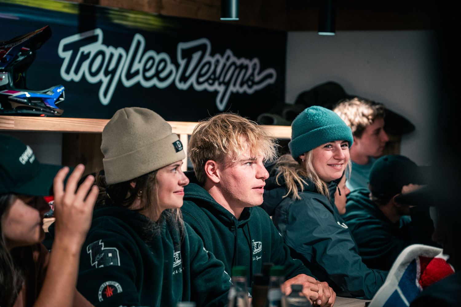 Athletes smile at a Troy Lee Designs event, wearing teal beanies and branded gear indoors.