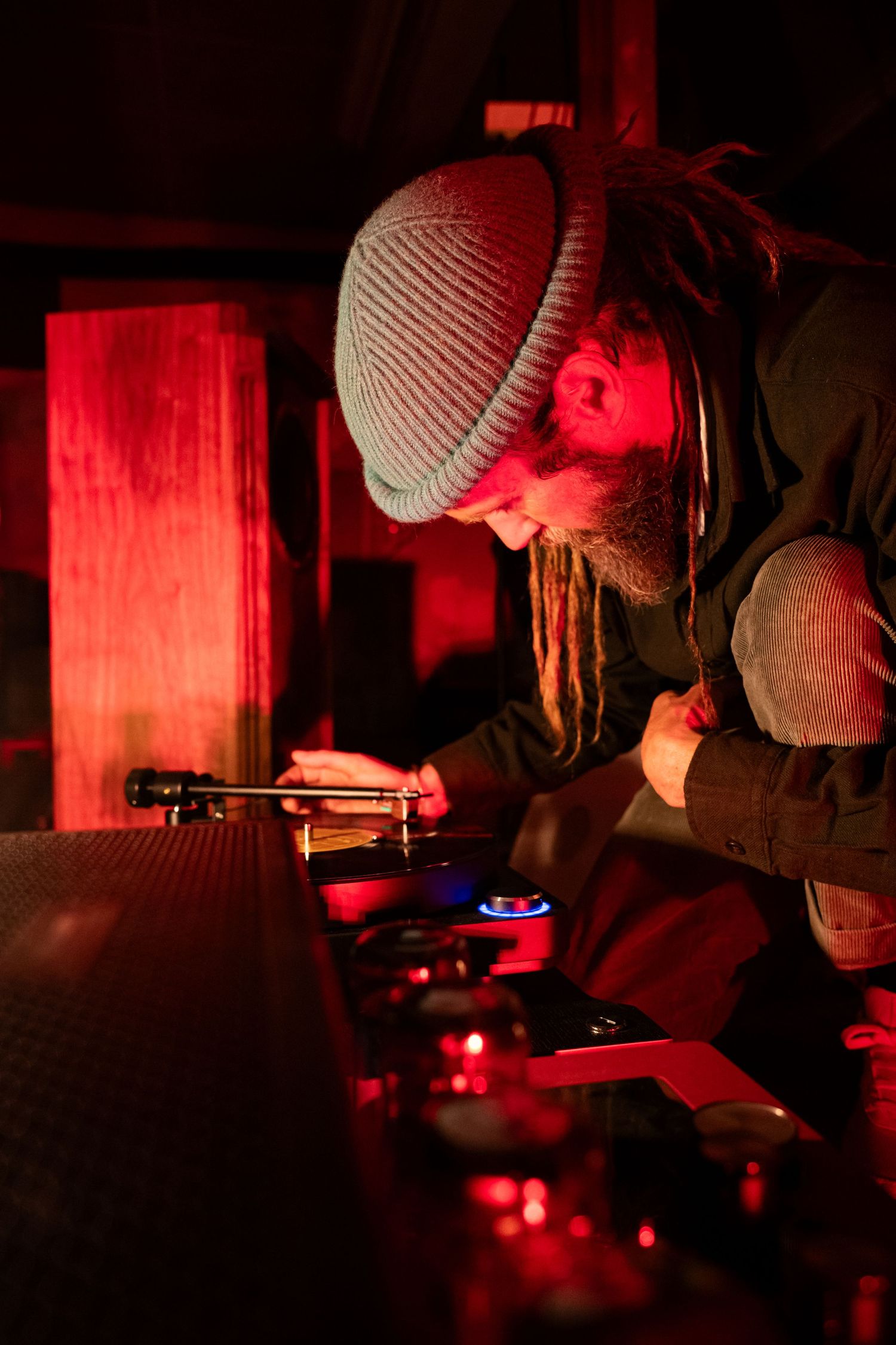 Man with dreadlocks and beanie hat leaning over a bar counter illuminated by red ambient lighting.
