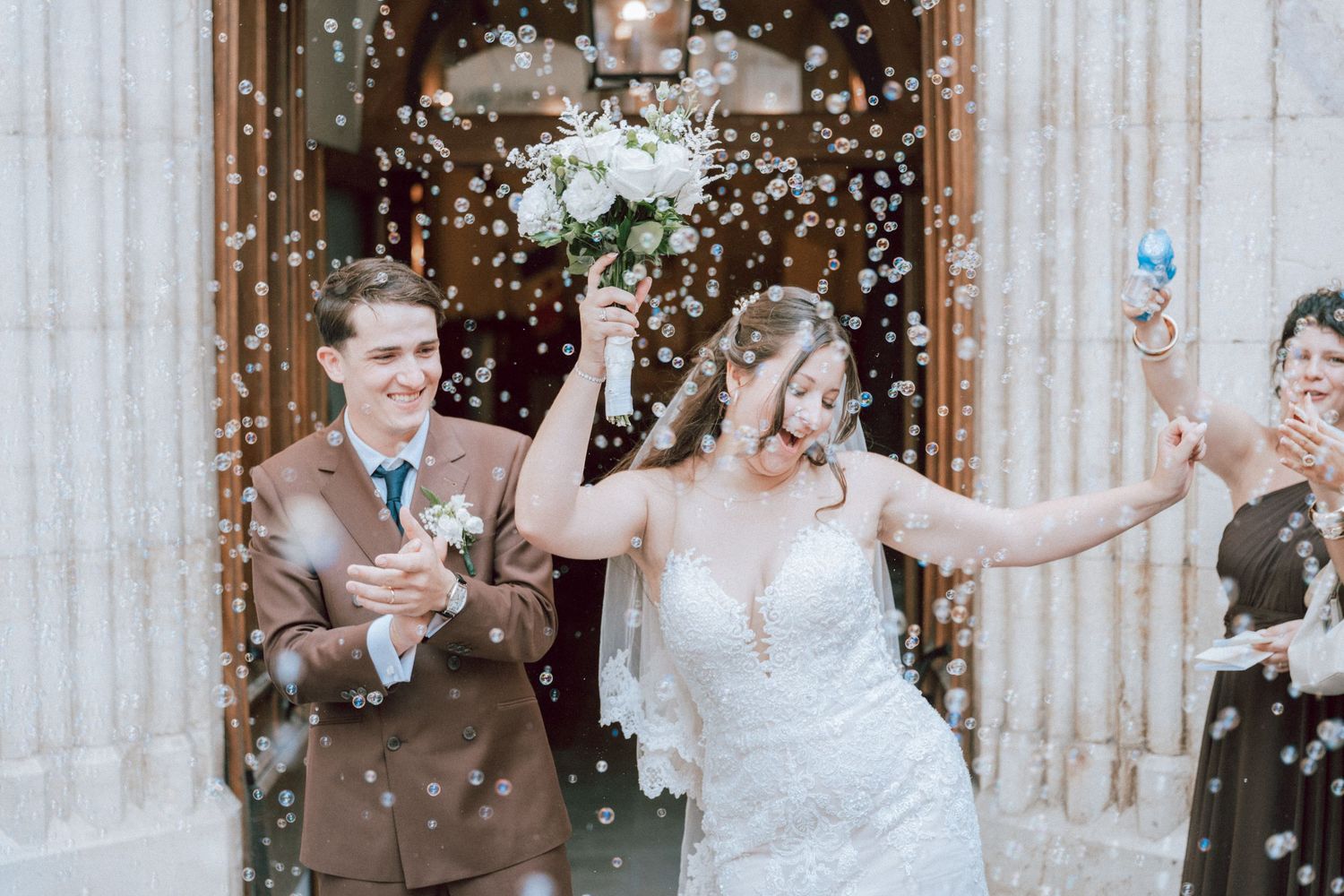 Joyful bride raising bouquet as couple exits church entrance surrounded by bubble confetti celebration.