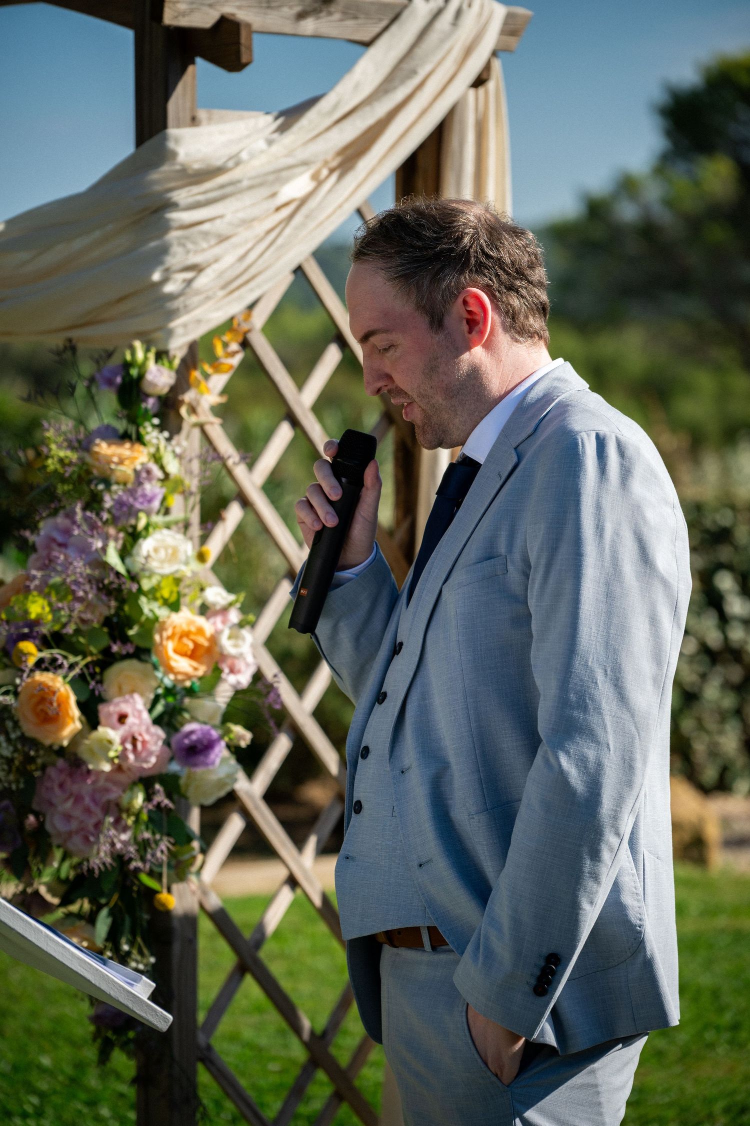 Man in light blue suit speaking into microphone at outdoor wedding ceremony with floral arch backdrop.