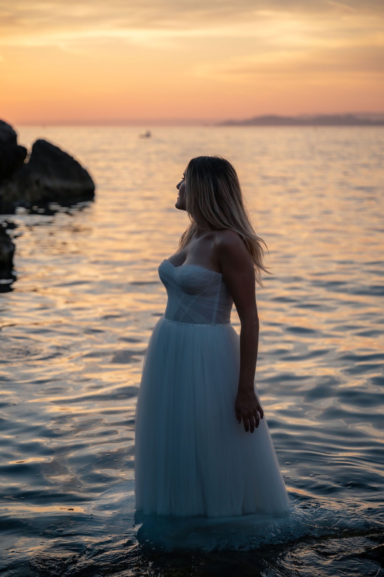 Bride in white strapless tulle gown standing in ocean water during golden sunset on rocky beach.