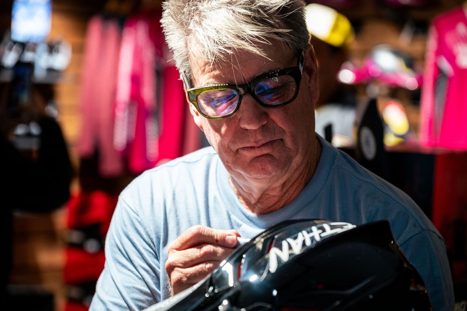 Man with gray hair and green glasses carefully signing a black helmet in a colorful workshop setting.