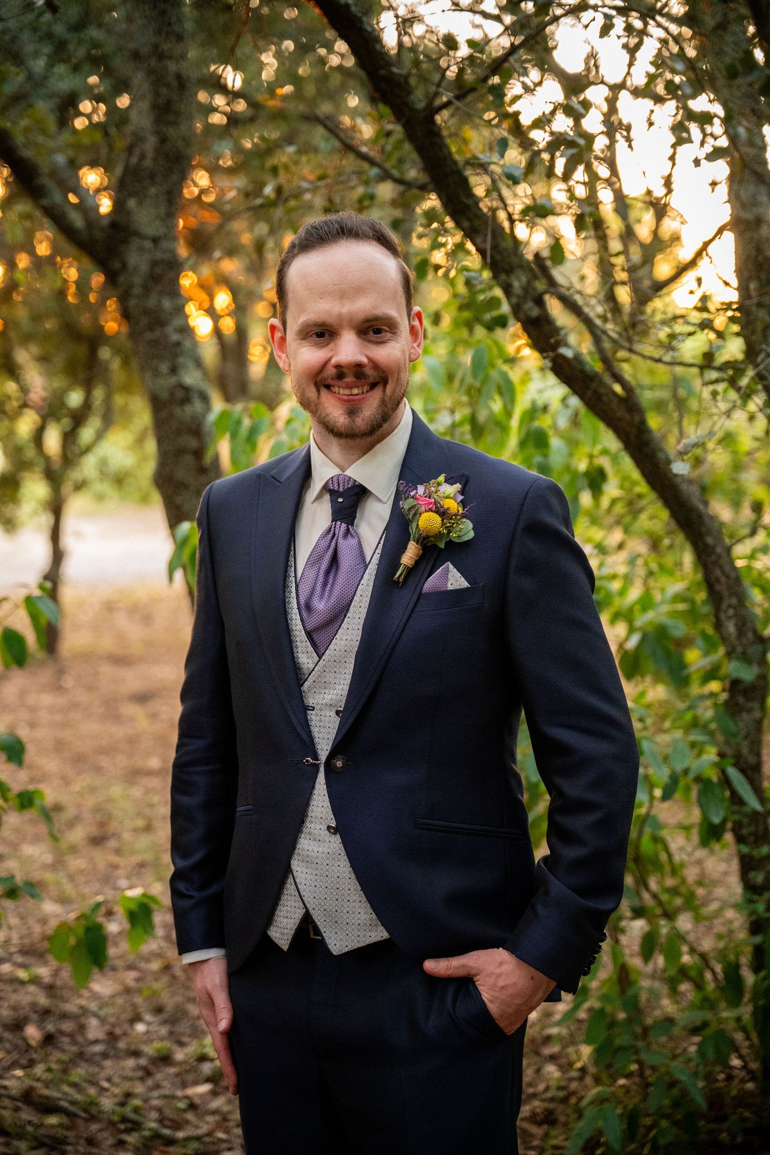 Smiling groom in navy suit with purple tie and floral boutonniere standing among trees at golden hour.