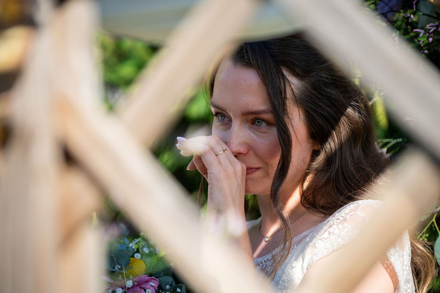 Emotional bride with dark hair covering her mouth with her hand, seen through a wooden lattice fence.
