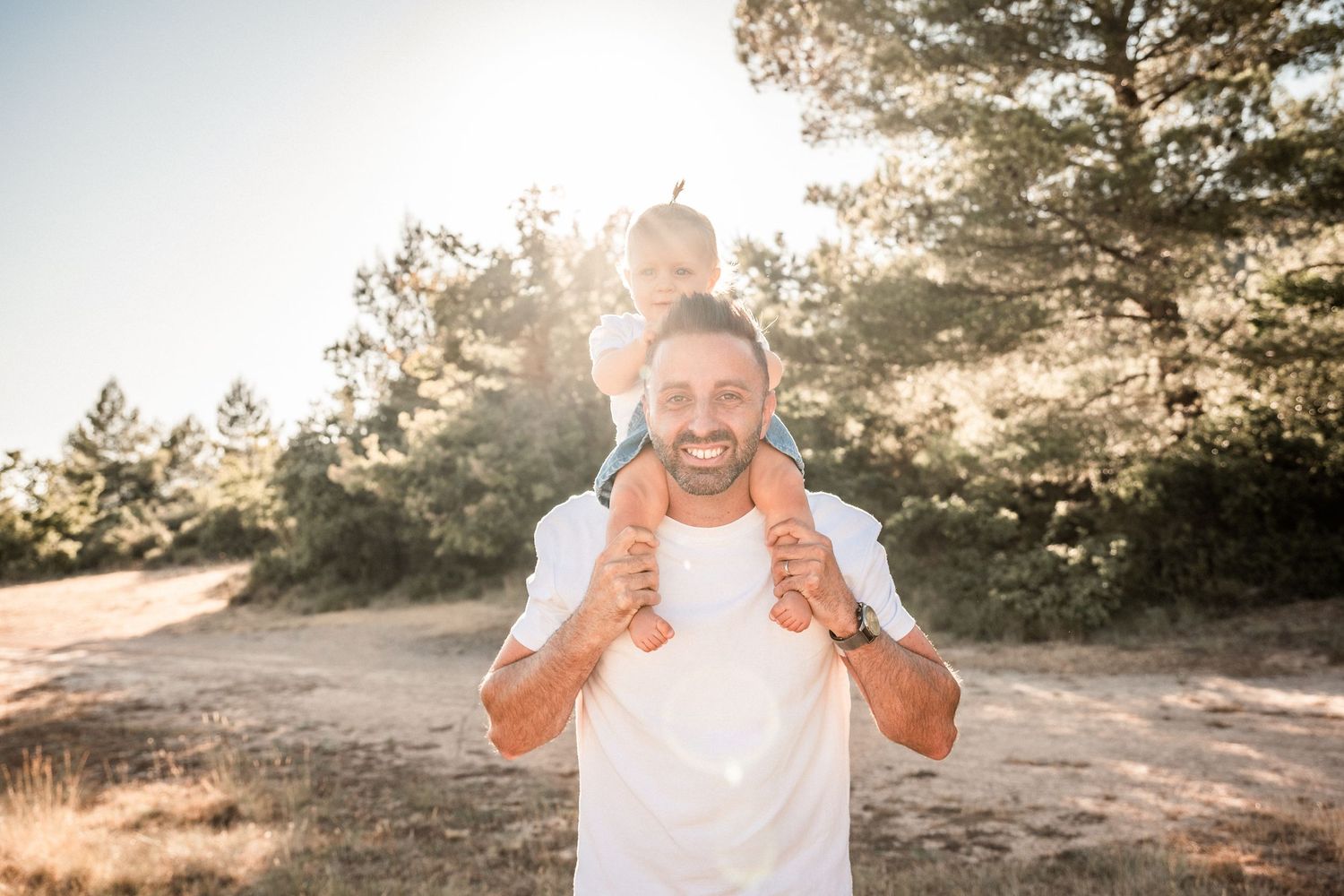 Happy father carrying young child on shoulders outdoors in sunny nature setting, smiling at camera.