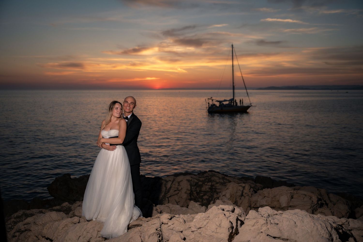 Newlyweds embracing on rocky coast at dramatic red sunset with sailboat.