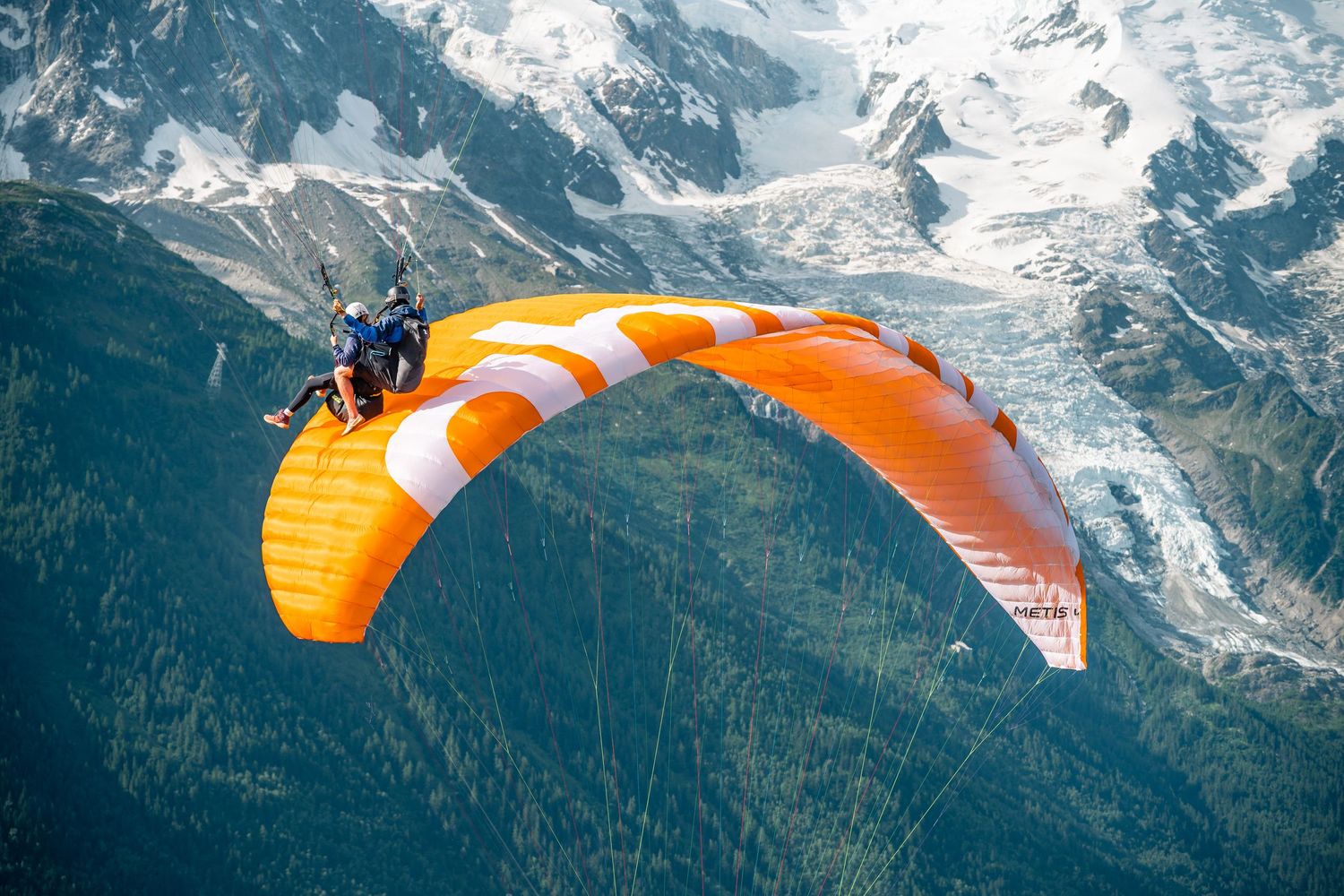 Orange tandem paraglider soaring above snow-capped Mont Blanc mountain range on clear day.
