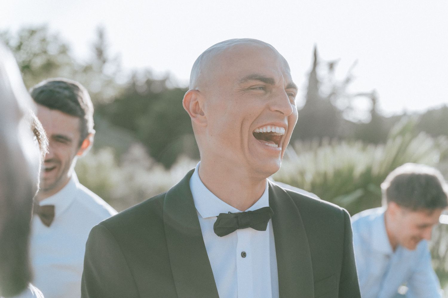 Bald groom in black tuxedo laughing joyfully with guests during sunny outdoor wedding celebration.