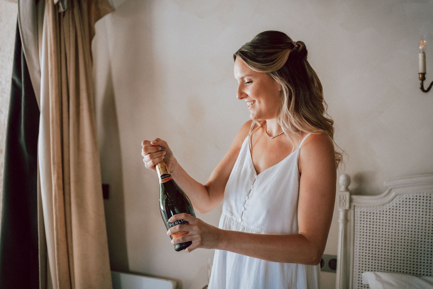 Bride in white dress opening champagne bottle while getting ready for wedding ceremony.