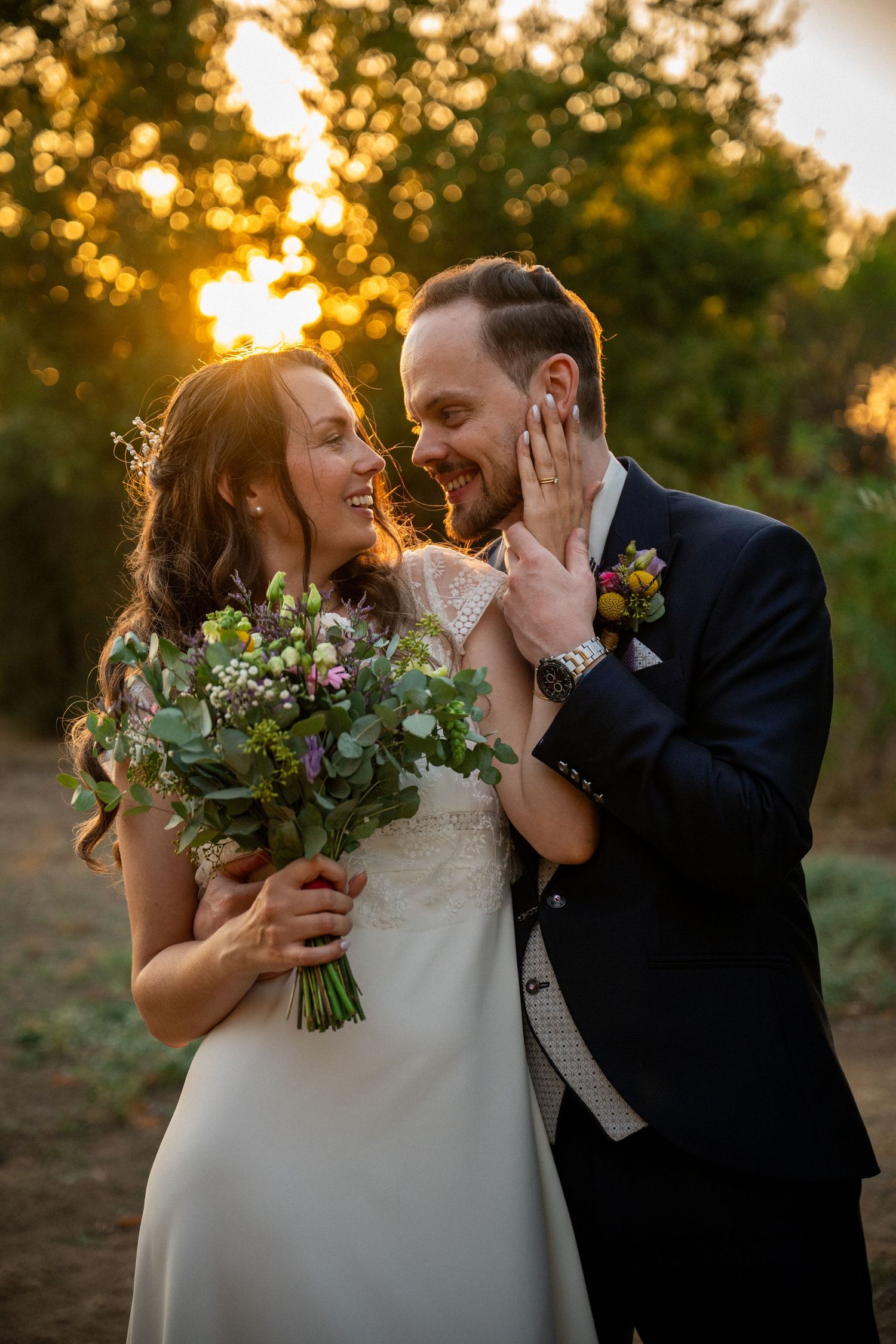 Smiling bride and groom sharing a tender moment at golden sunset, bride holding wildflower bouquet.
