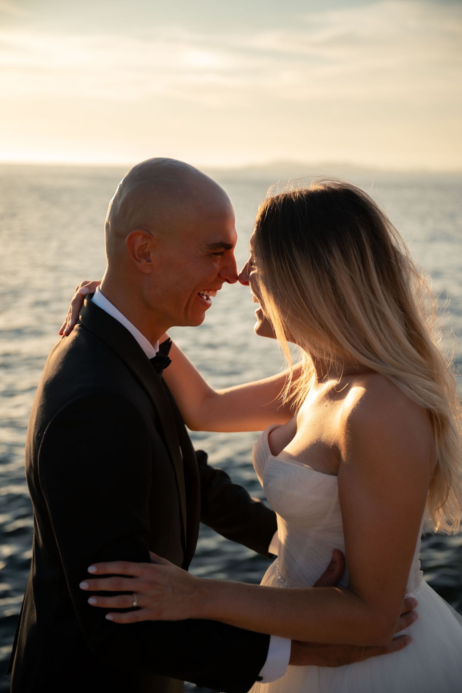 Bride and groom embrace at sunset by the ocean, smiling at each other in wedding attire.