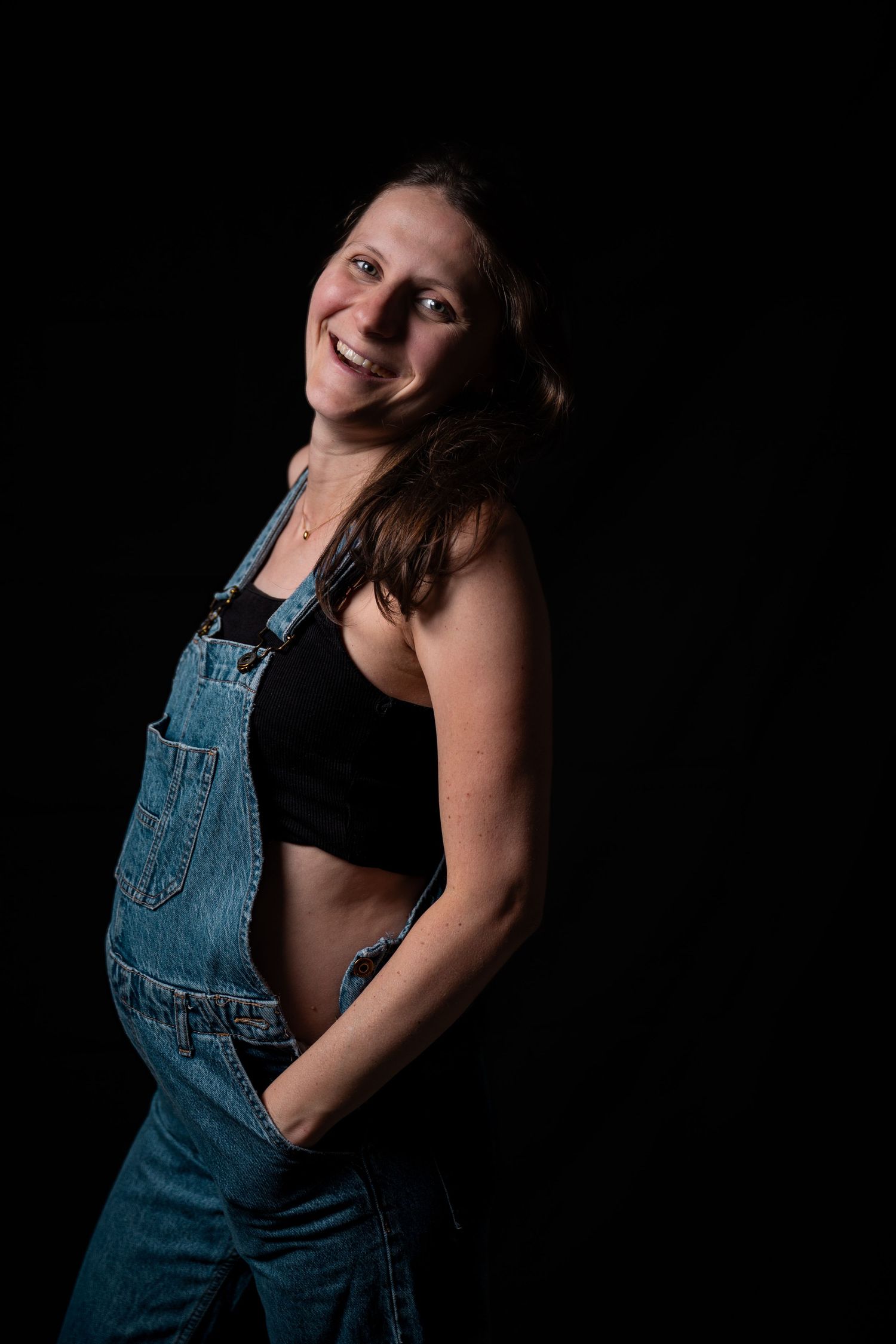 Smiling pregnant woman in denim overalls and black crop top posing against a dark background.
