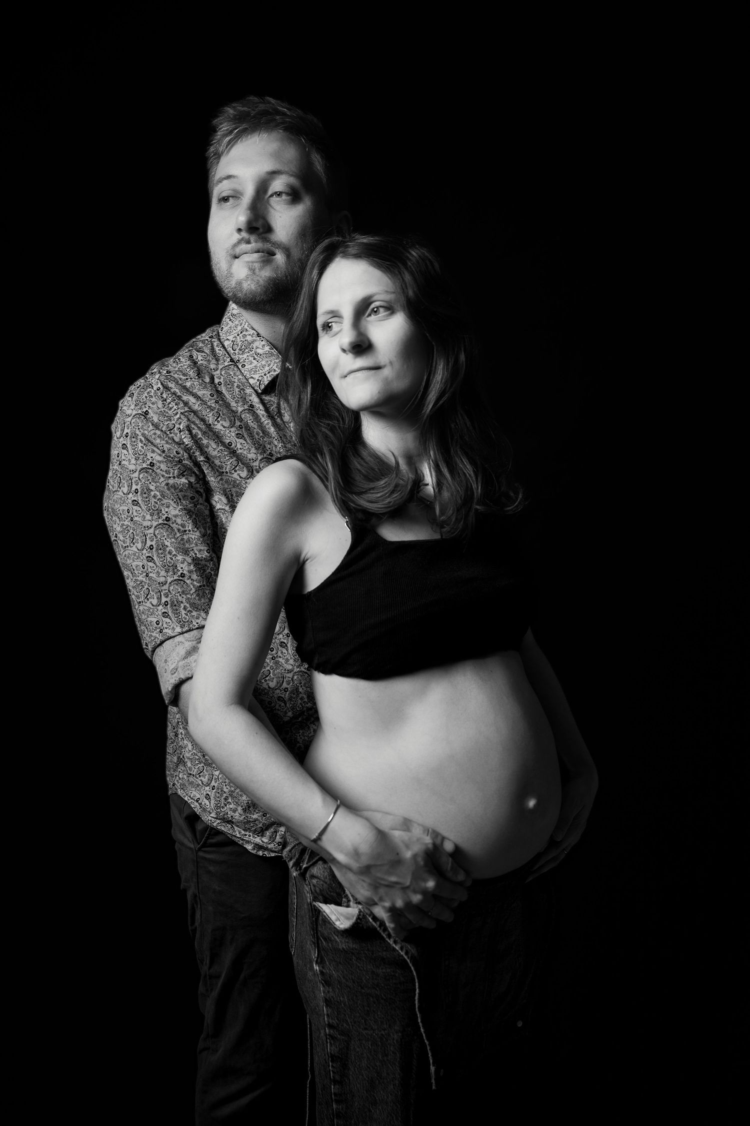 Black and white maternity portrait of a couple embracing, showing pregnant belly, against a dark studio background.