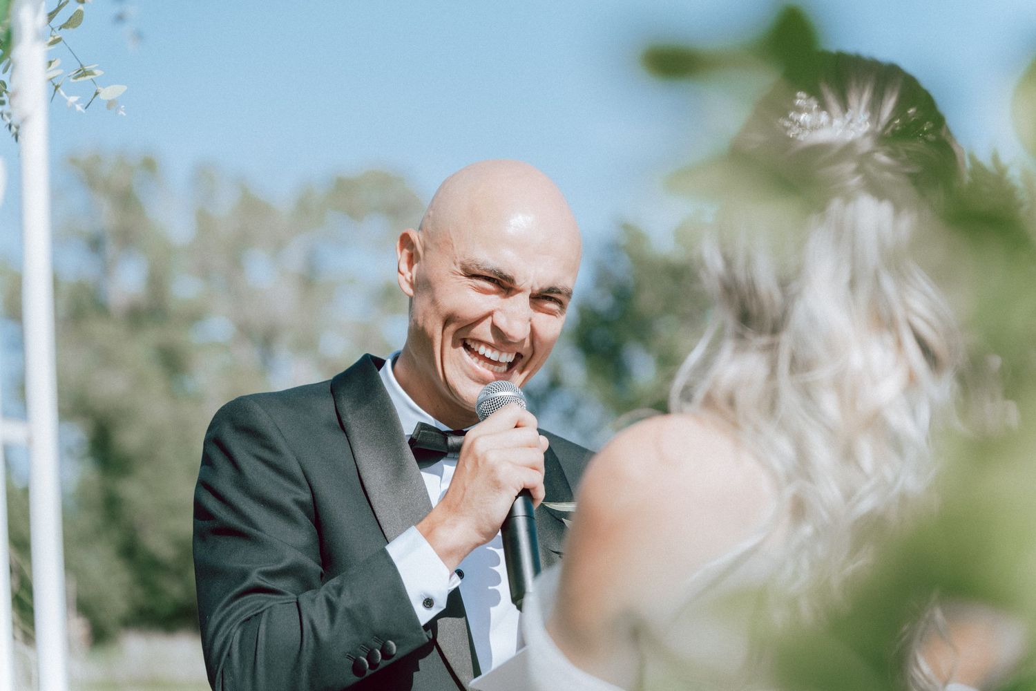 Bald groom in dark tuxedo smiles joyfully at bride during outdoor wedding ceremony.
