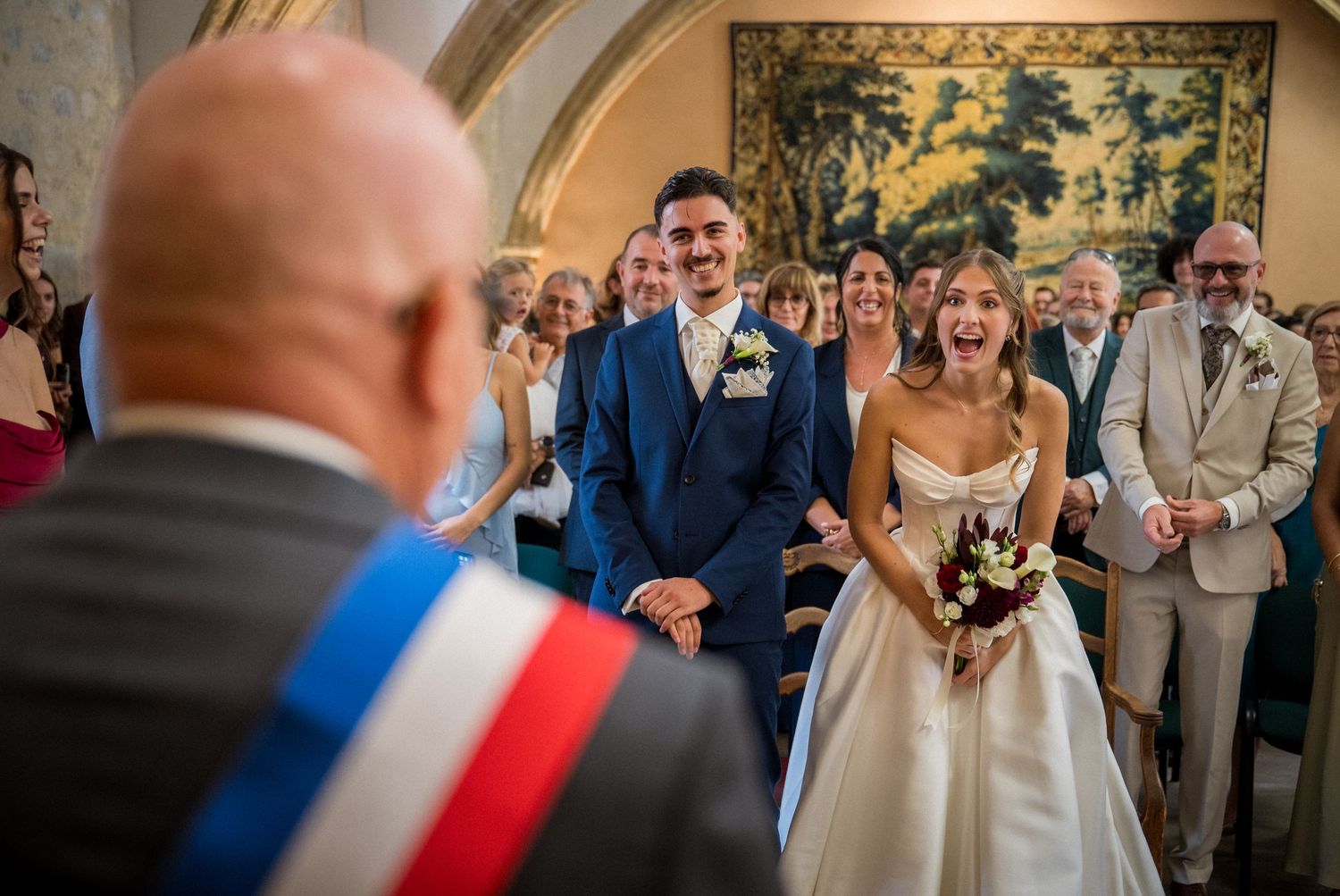 Bride and groom laughing joyfully during civil wedding ceremony with guests and official wearing French tricolor sash.