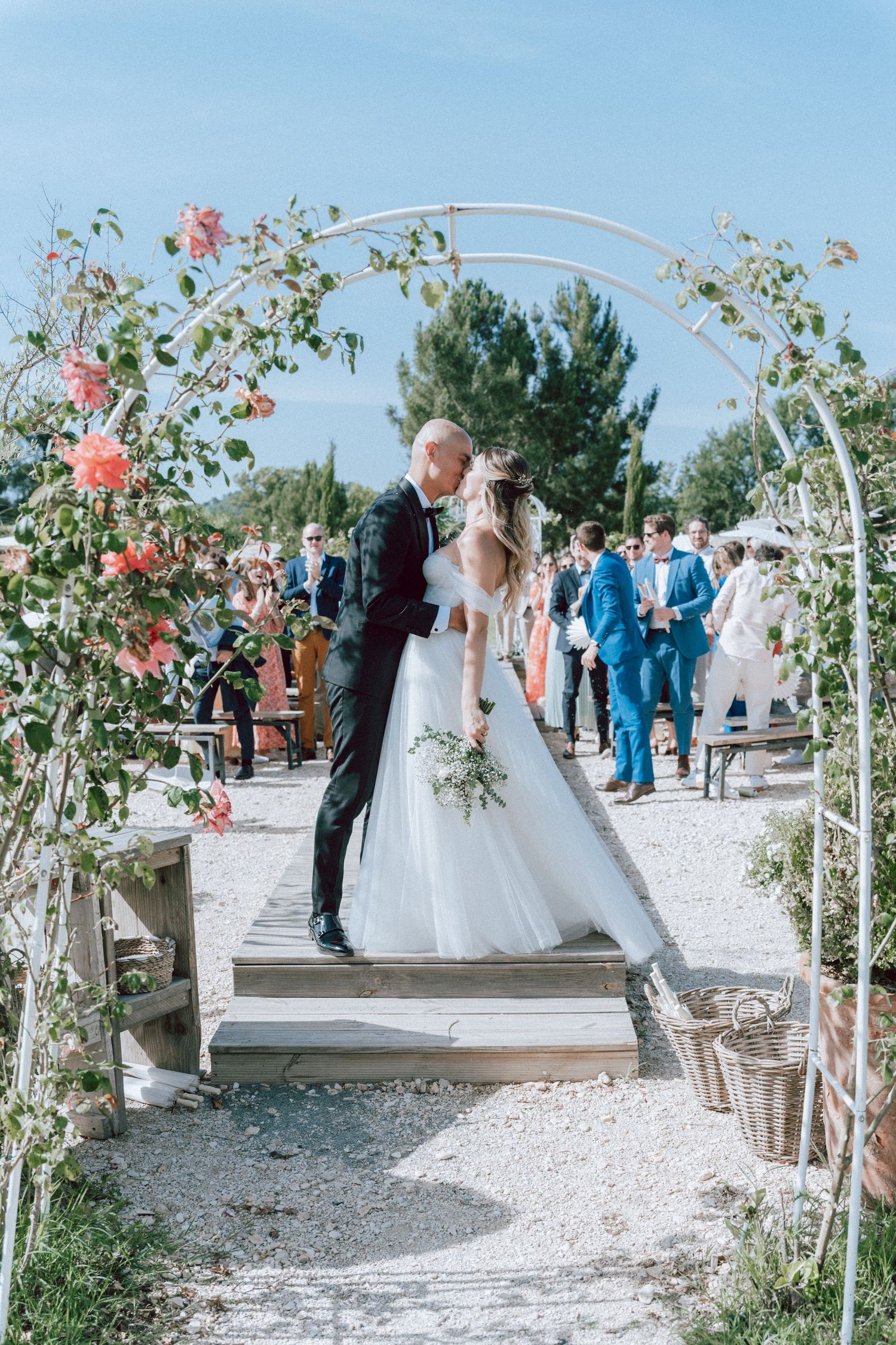 Bride and groom share a first kiss under a floral arch at an outdoor wedding ceremony with guests in background.