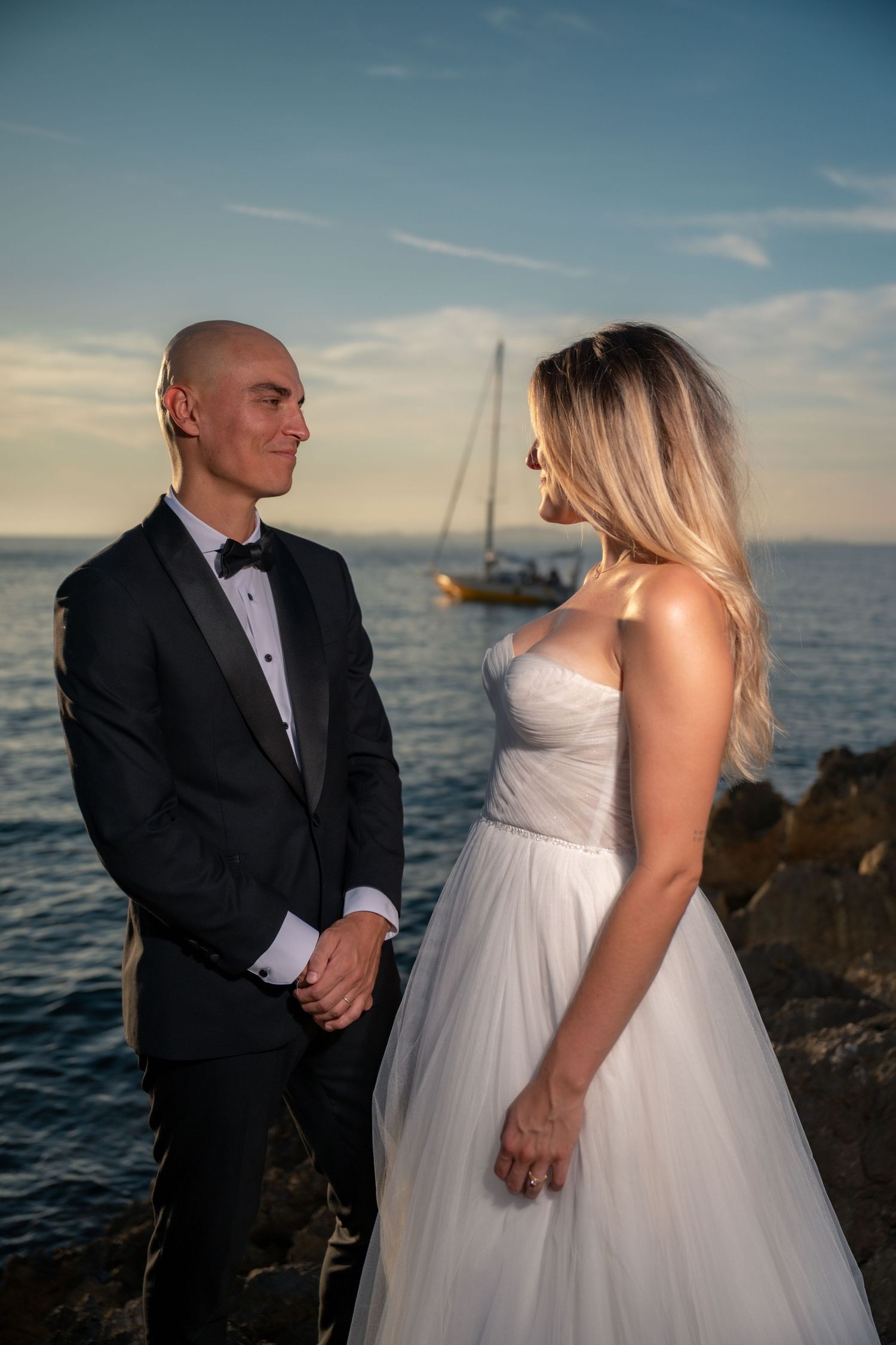 Bride and groom share a romantic moment by the sea at sunset, with a sailboat in the background.