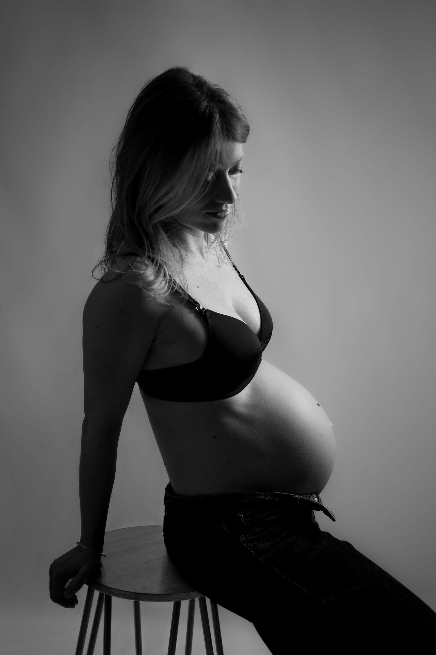 Black and white silhouette of a pregnant woman sitting on a stool wearing a black bra and jeans.