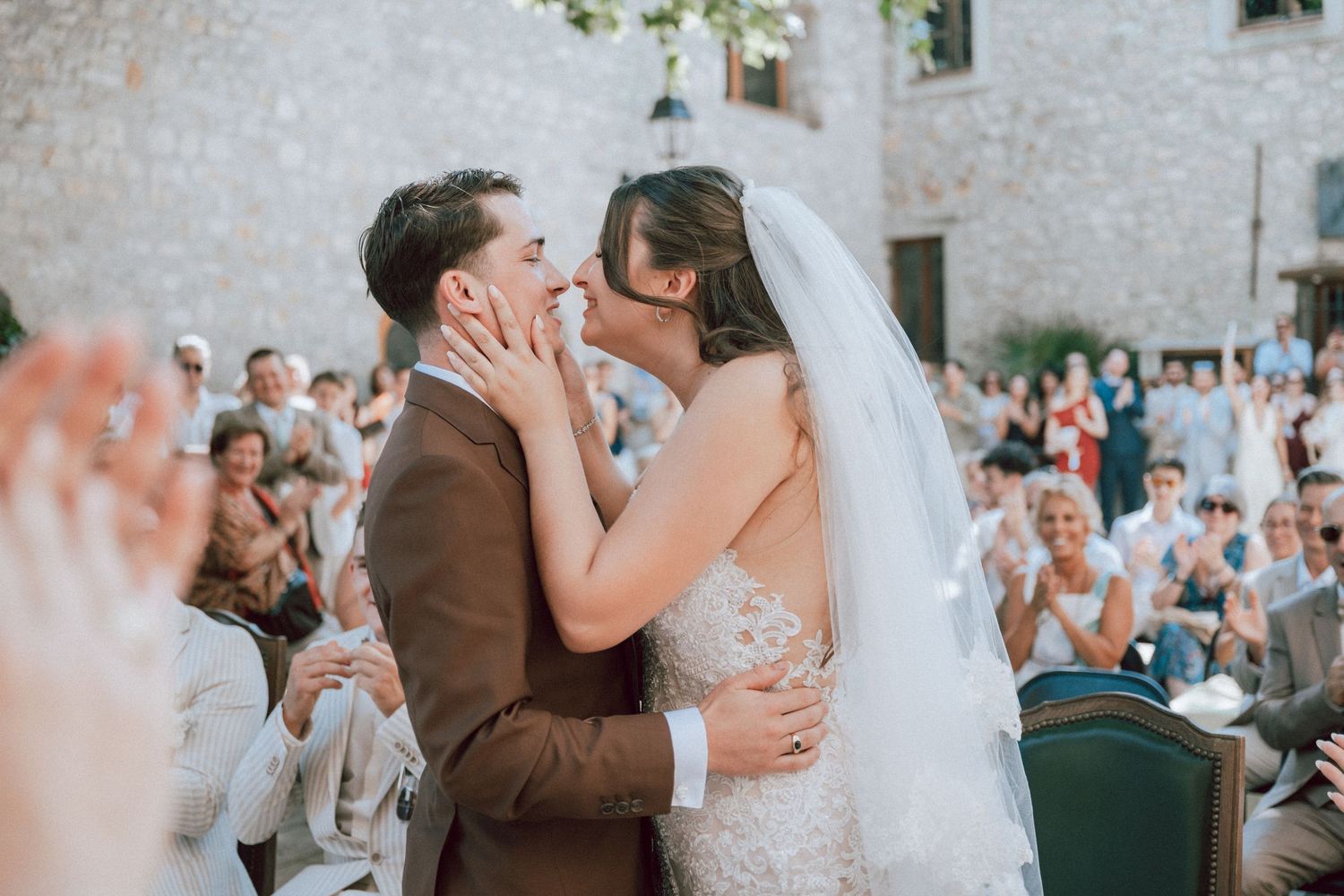 Newlyweds share first kiss at wedding ceremony as guests applaud in rustic stone venue.