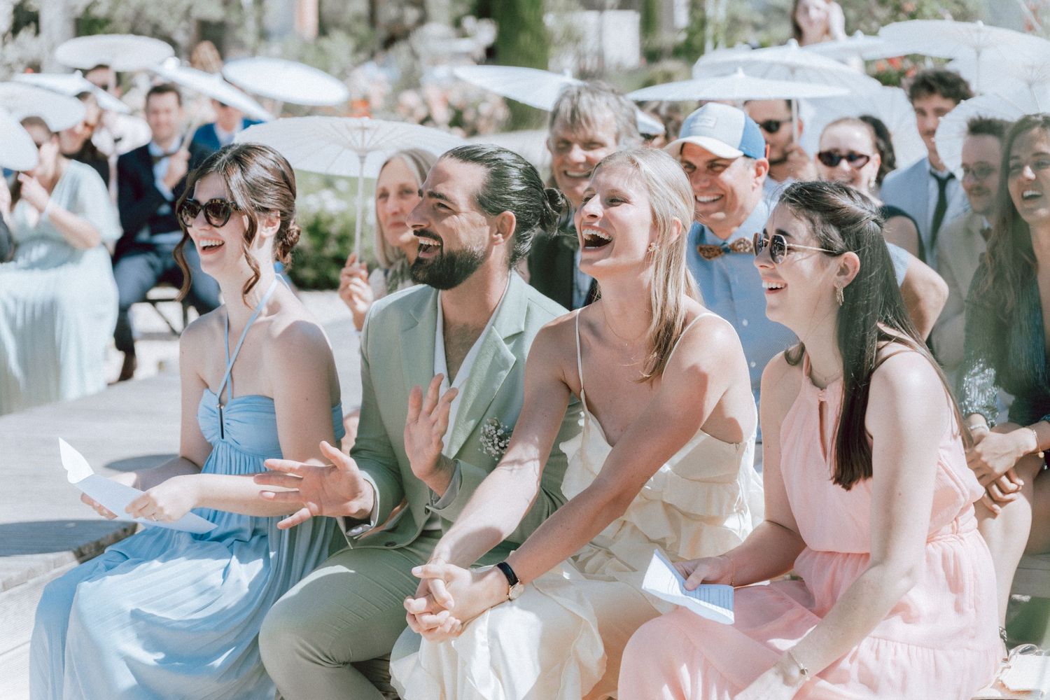 Wedding guests laughing and reacting joyfully while seated at sunny outdoor wedding ceremony.