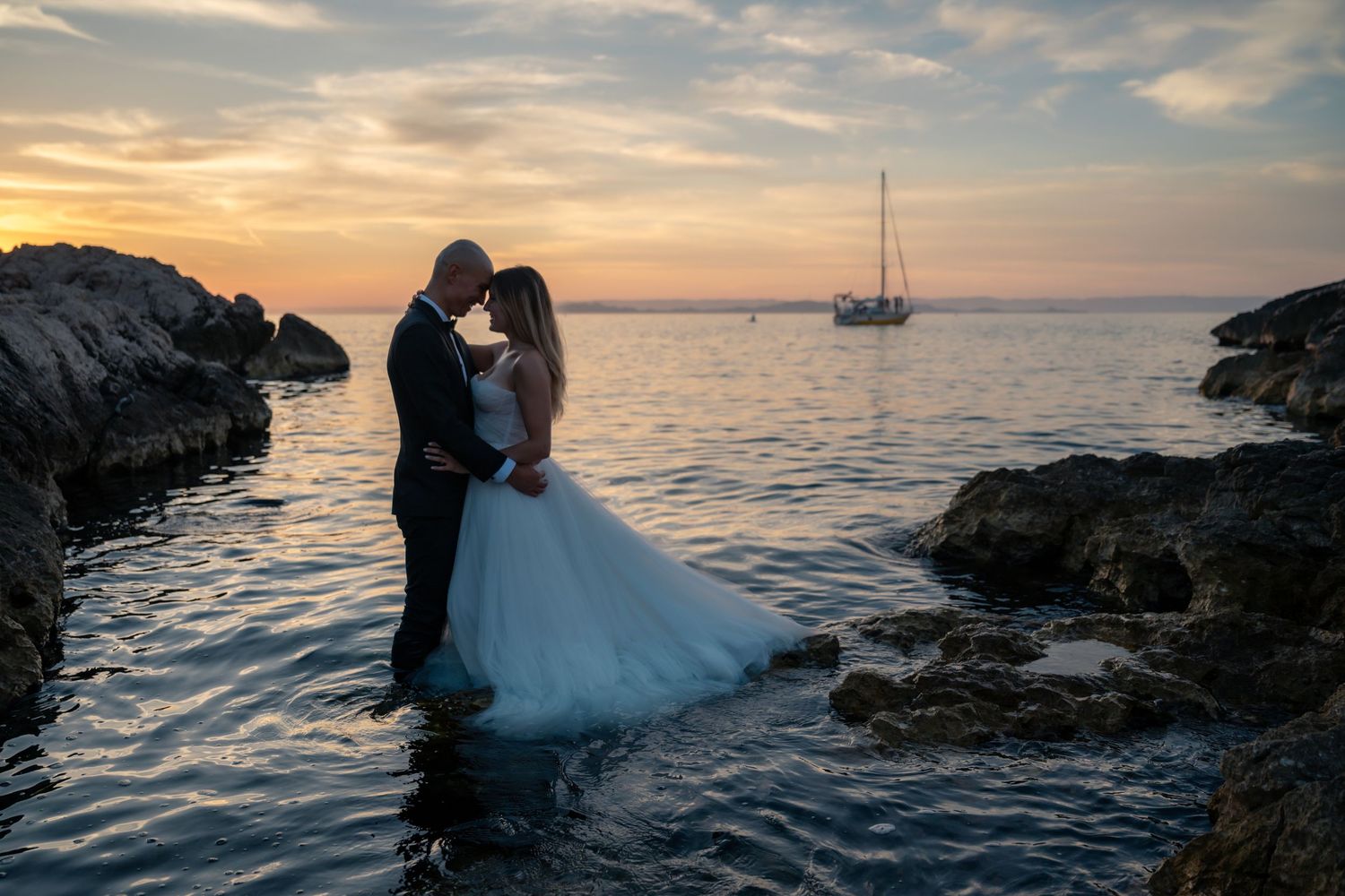 Newlyweds share a kiss standing in shallow water by rocks at sunset with sailboat behind.