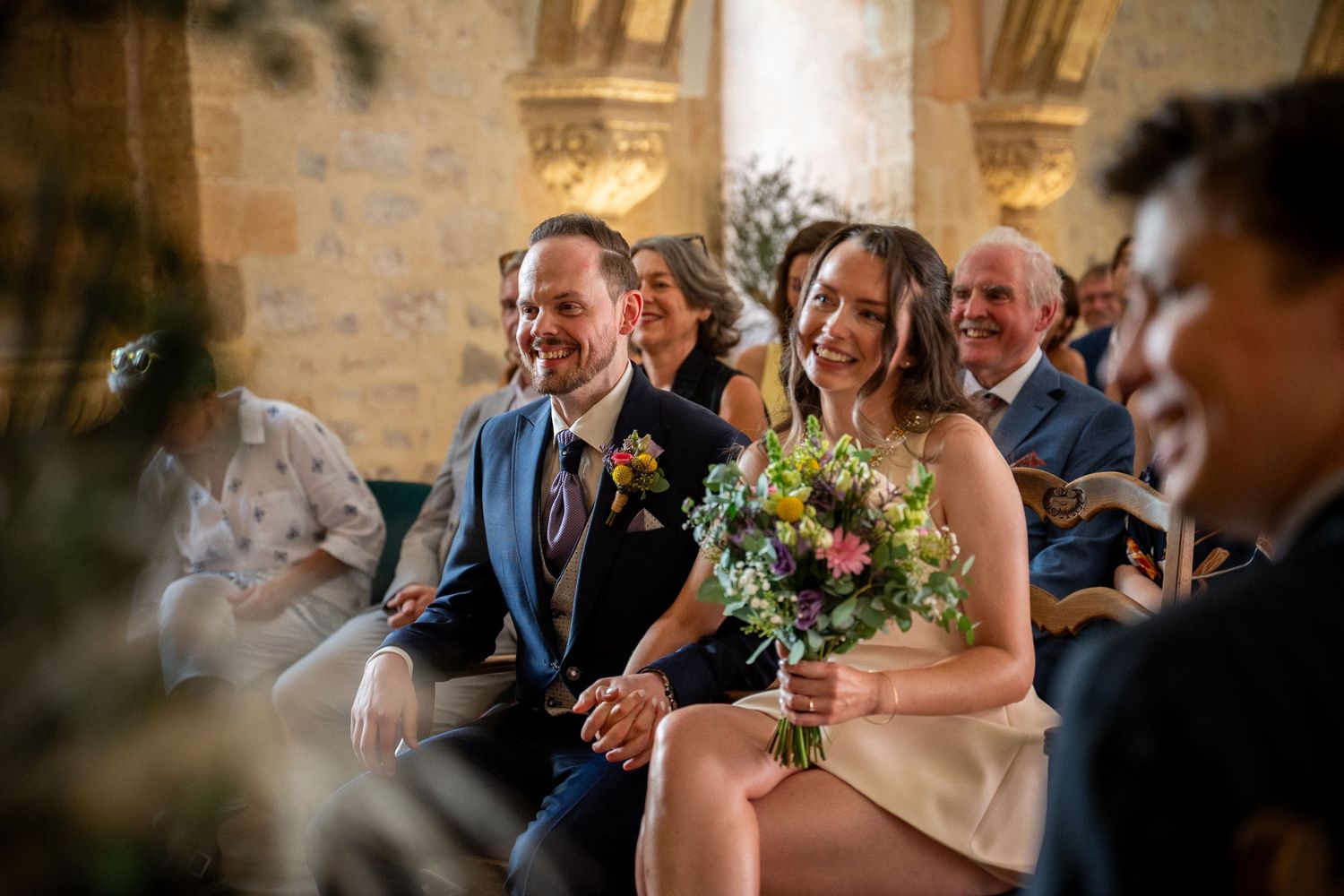 Happy couple sitting together during wedding ceremony, bride holding colorful bouquet, groom in blue suit, guests smiling behind them.