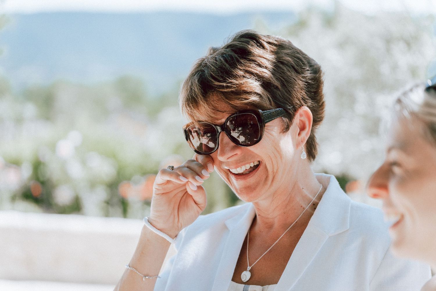 Smiling woman with short dark hair wearing sunglasses and white blazer outdoors at a sunny wedding event.