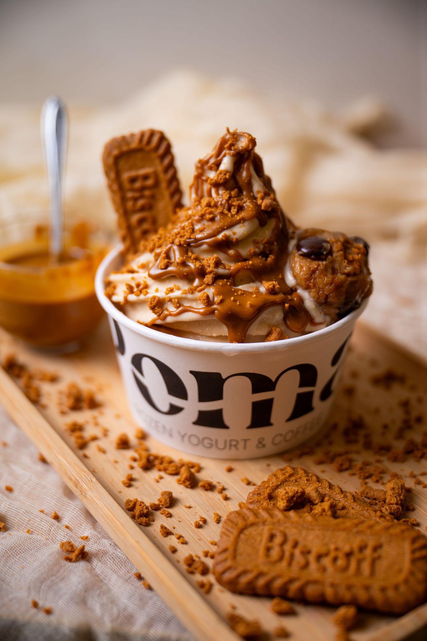 Foma frozen yogurt cup topped with caramel sauce, biscoff cookies, and crumbles on a wooden board.
