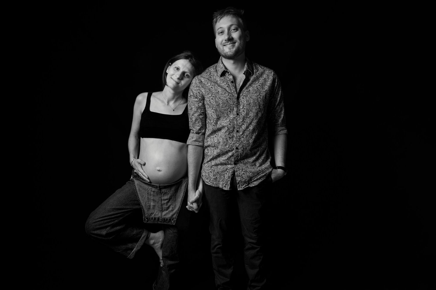 Black and white maternity photo of pregnant woman and partner holding hands against dark background.