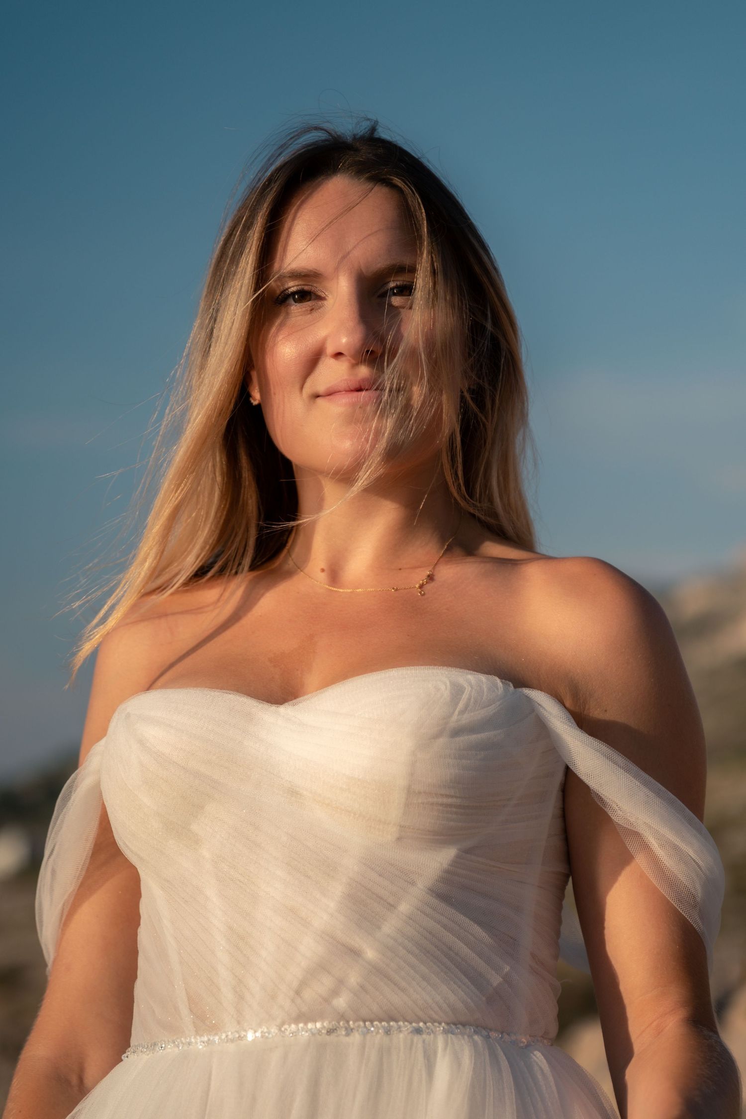Beautiful bride in white off-shoulder wedding gown posing outdoors under blue sky.