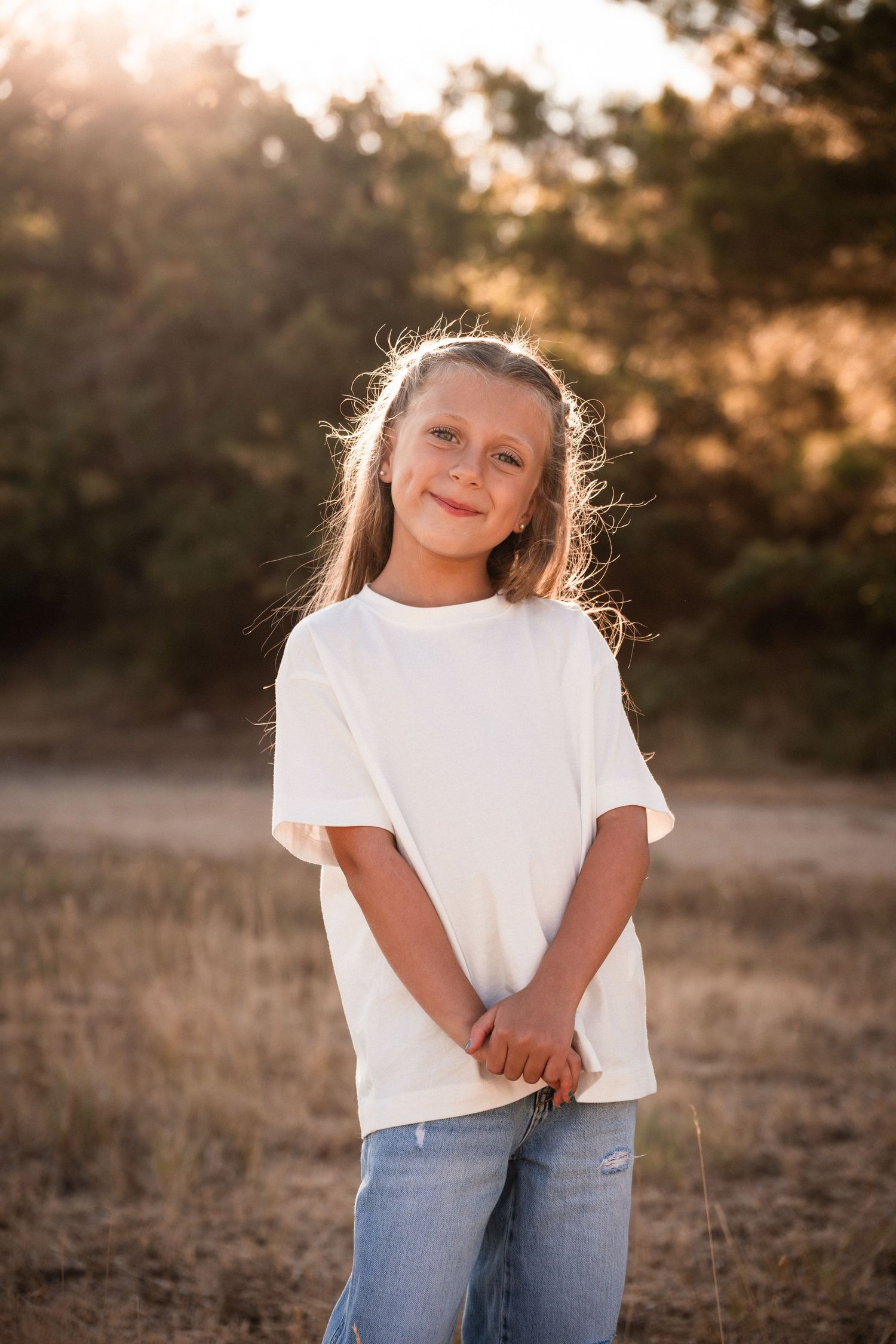 Young girl in white t-shirt and jeans smiling outdoors in golden sunset light with trees in background.