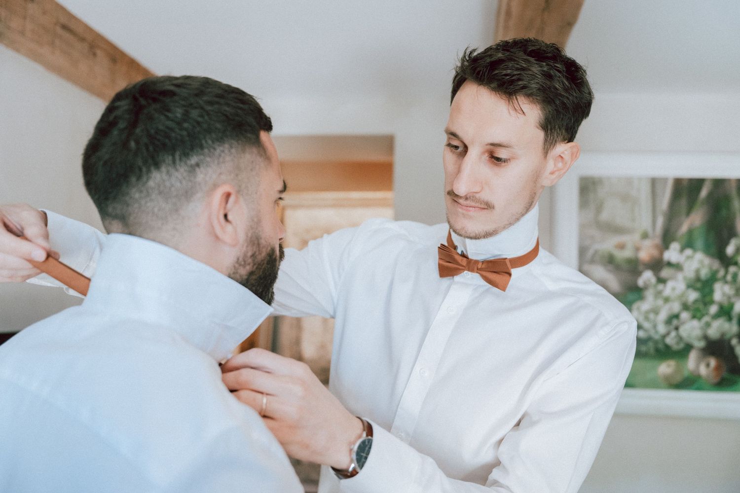 Two men in white shirts, one wearing a brown bow tie, helping the other prepare for a wedding ceremony.