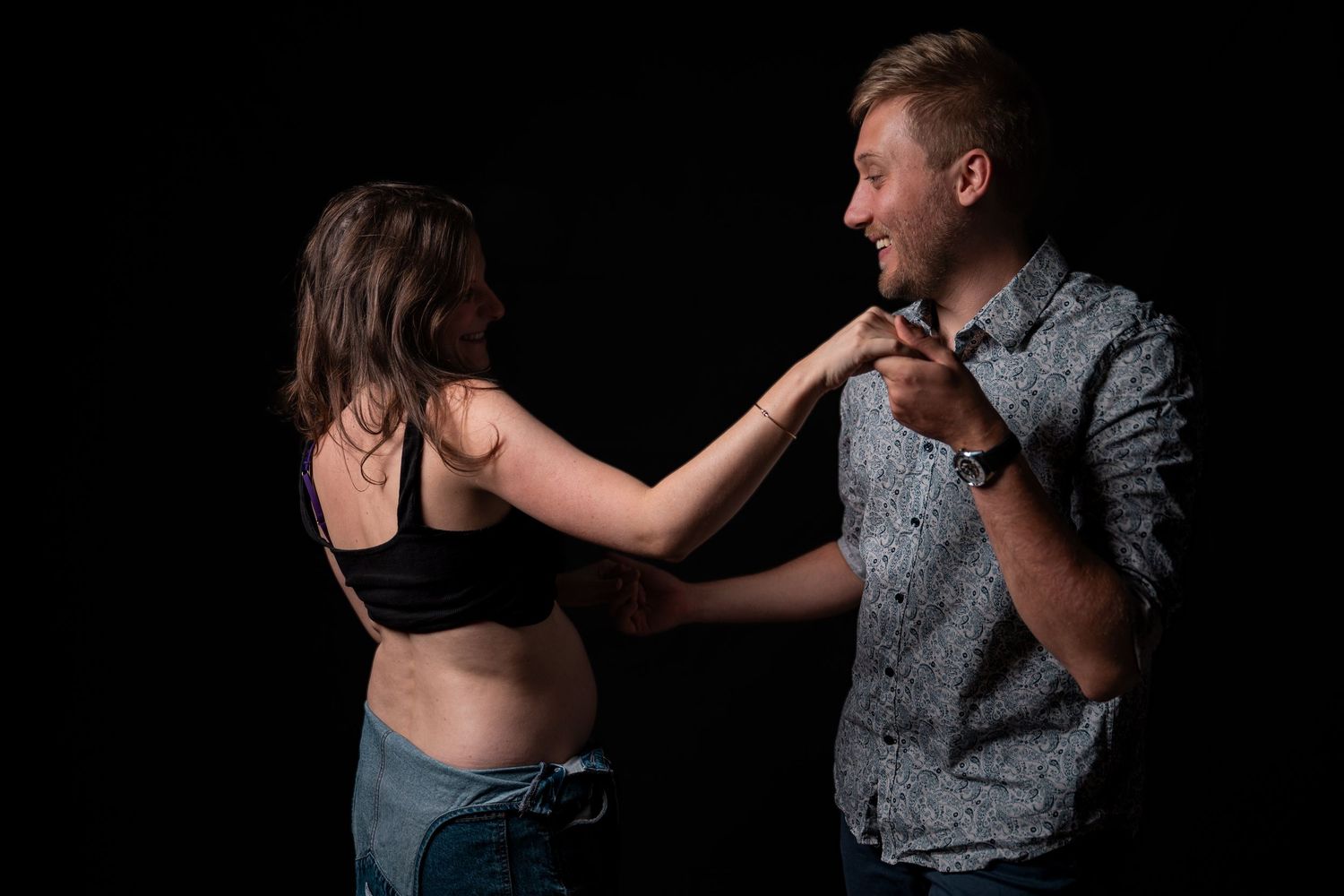 A man and woman facing each other, she touches his tie while he smiles, against a dark background.
