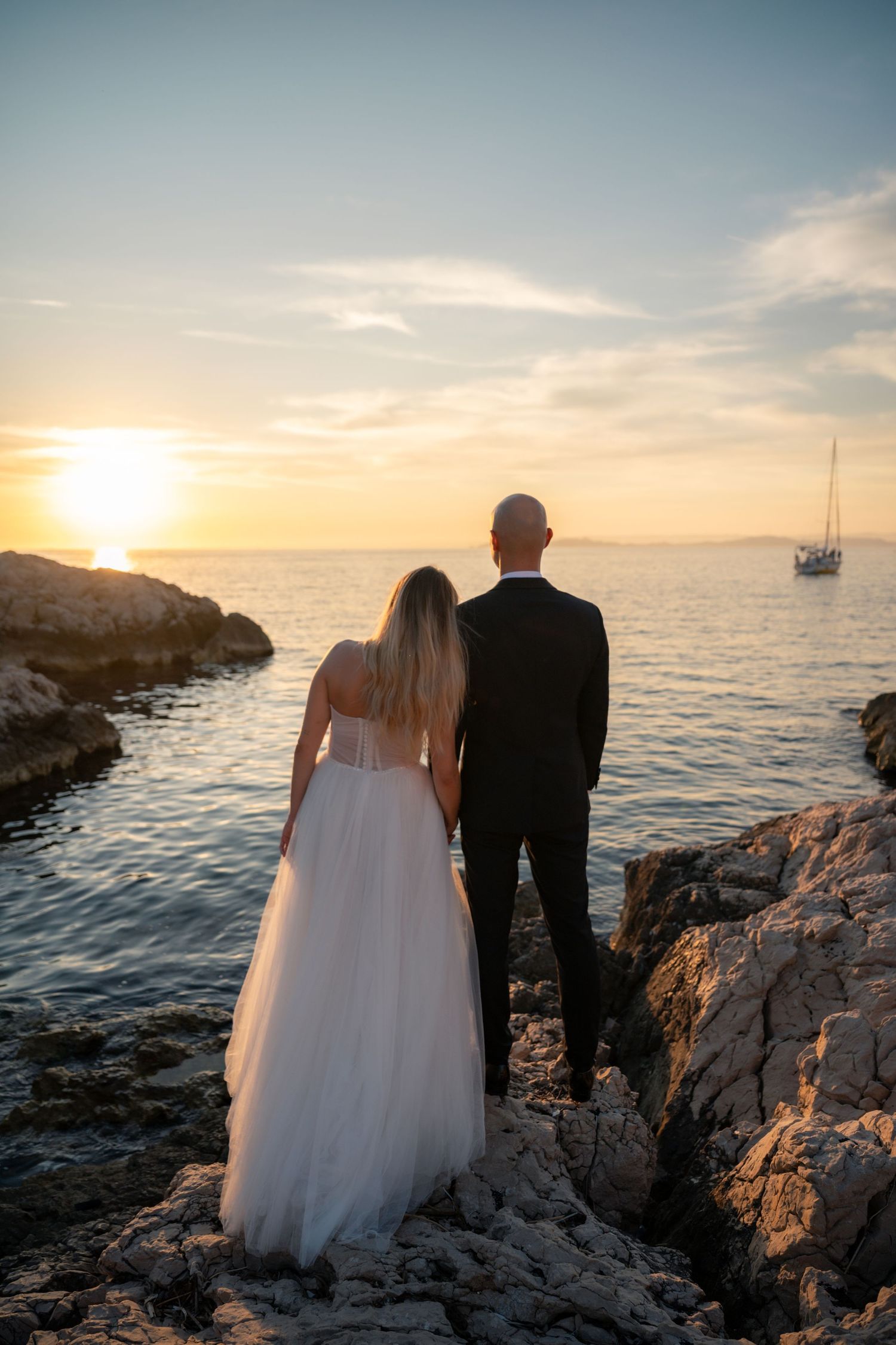 Bride and groom standing on rocky shore watching golden sunset over calm ocean with sailboat in distance.